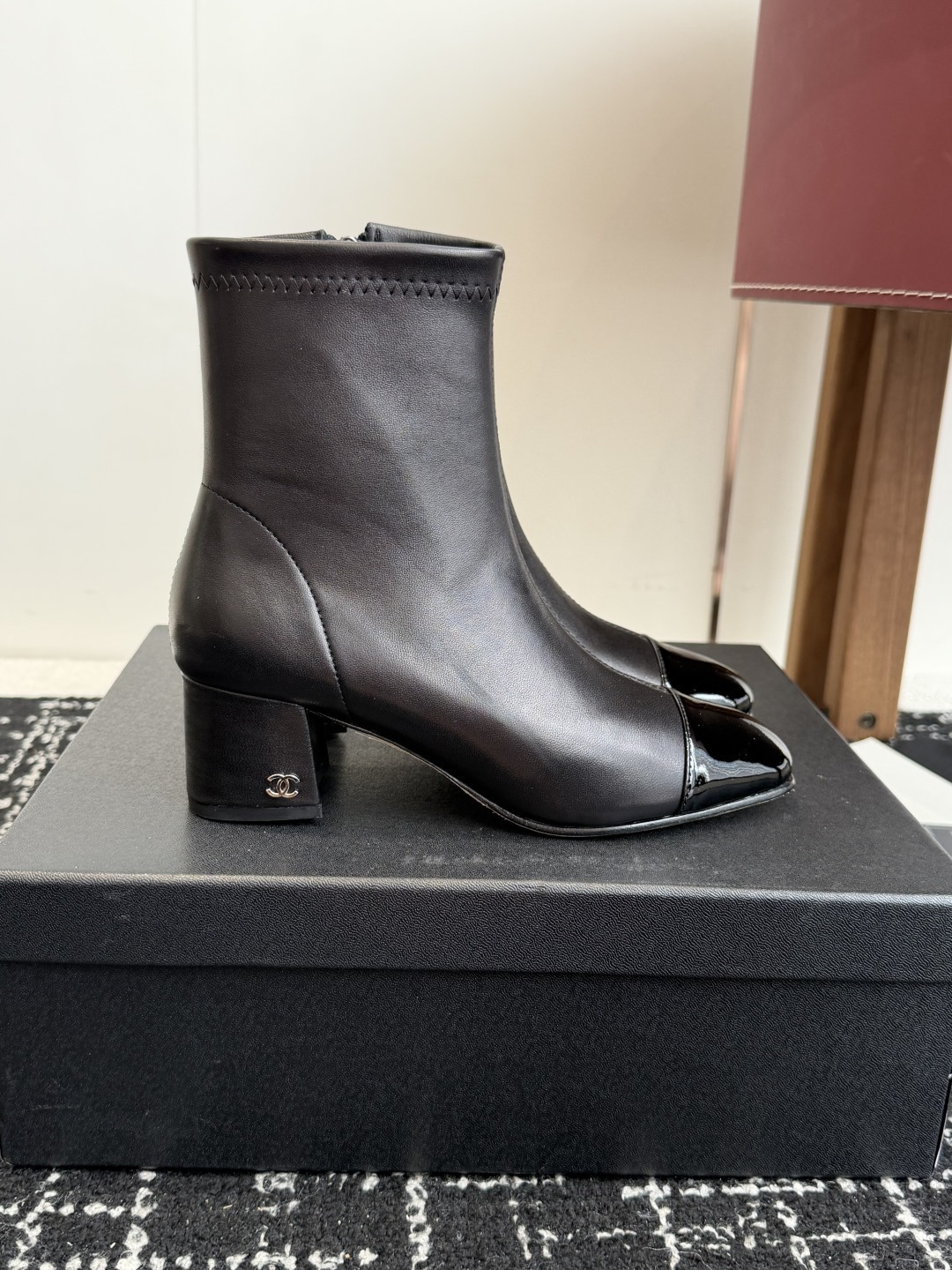 A pair of black Chanel leather ankle boots with glossy patent leather toe caps and block heels featuring the CC logo, displayed on a black box. - Hình 2