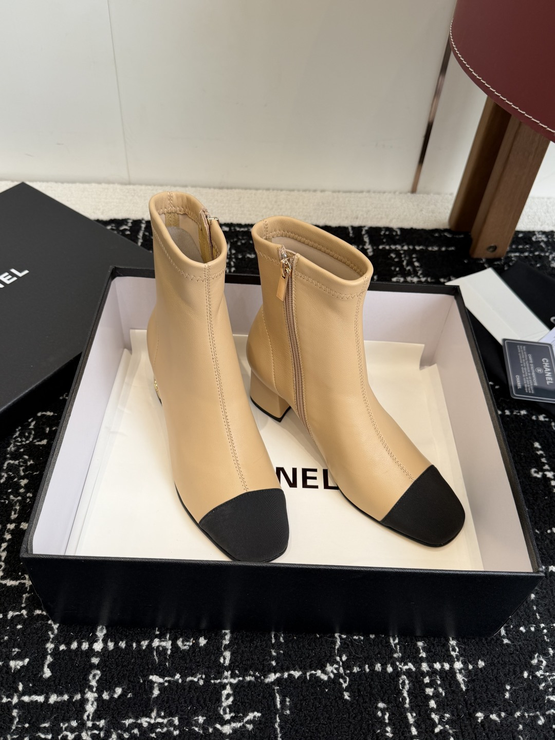 A pair of Chanel two-tone beige leather ankle boots with black fabric cap-toes and gold CC logo on the block heels, presented on a black Chanel box. - Hình 3