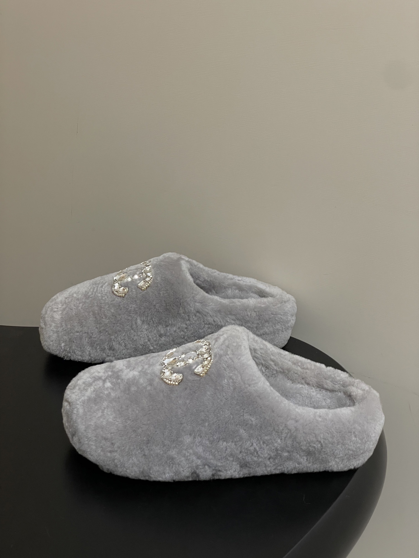 A pair of grey plush faux fur slippers with a sparkling rhinestone crystal logo on a black surface. - Hình 3
