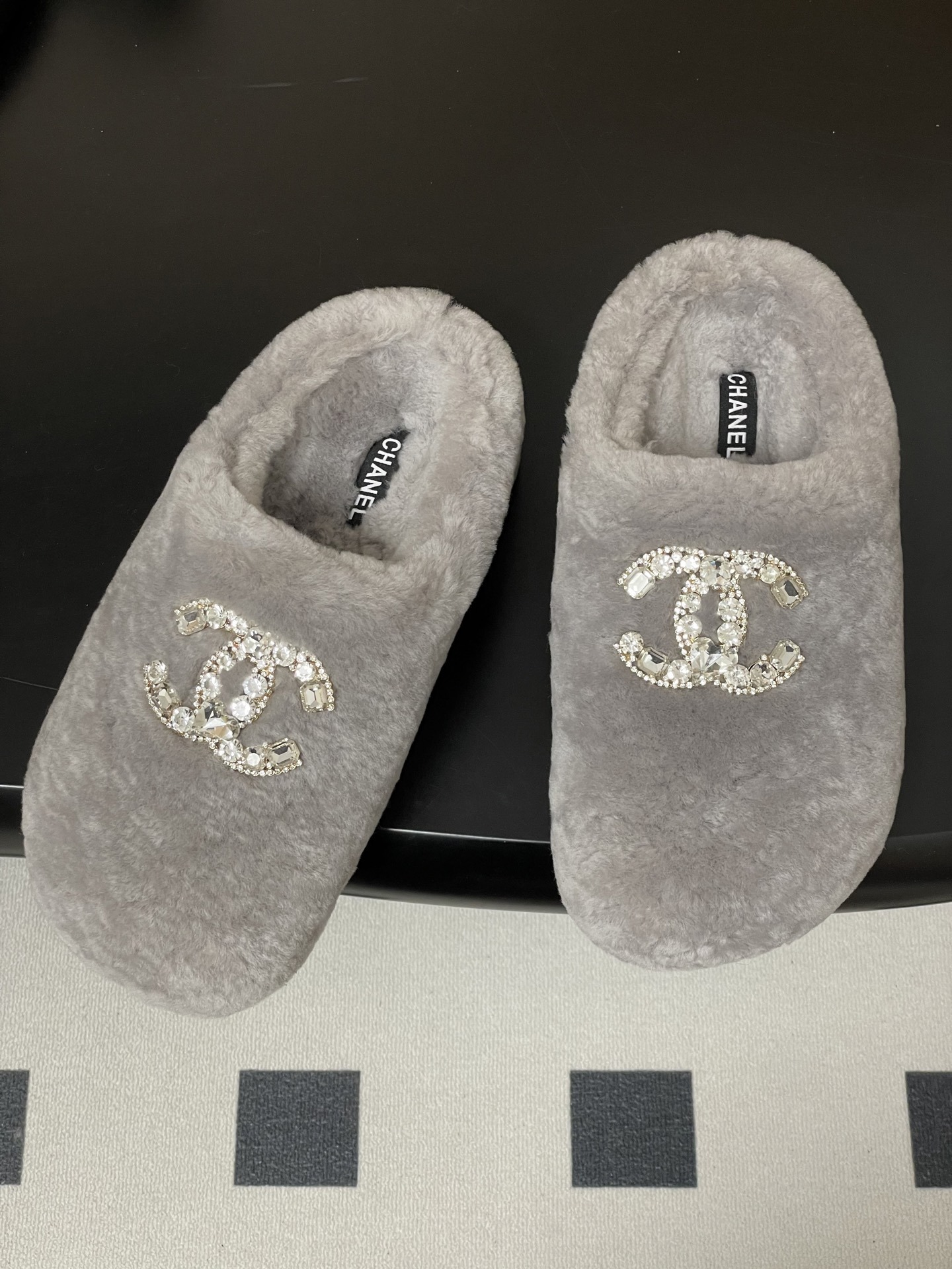 A pair of grey plush faux fur slippers with a sparkling rhinestone crystal logo on a black surface. - Hình 2