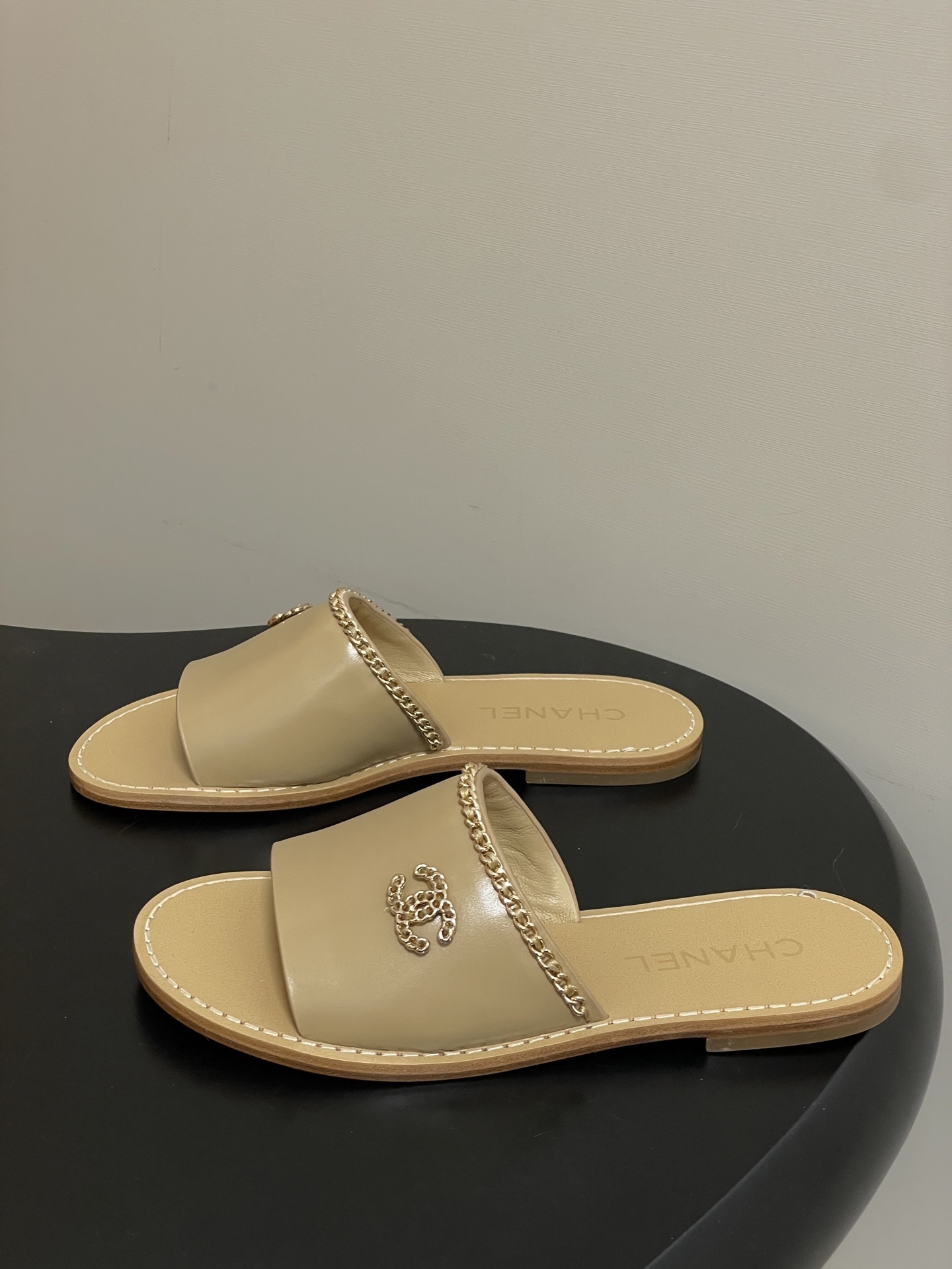 Luxury beige Chanel leather flat sandals with gold chain trim and interlocking CC logo on a black surface. - Hình 3