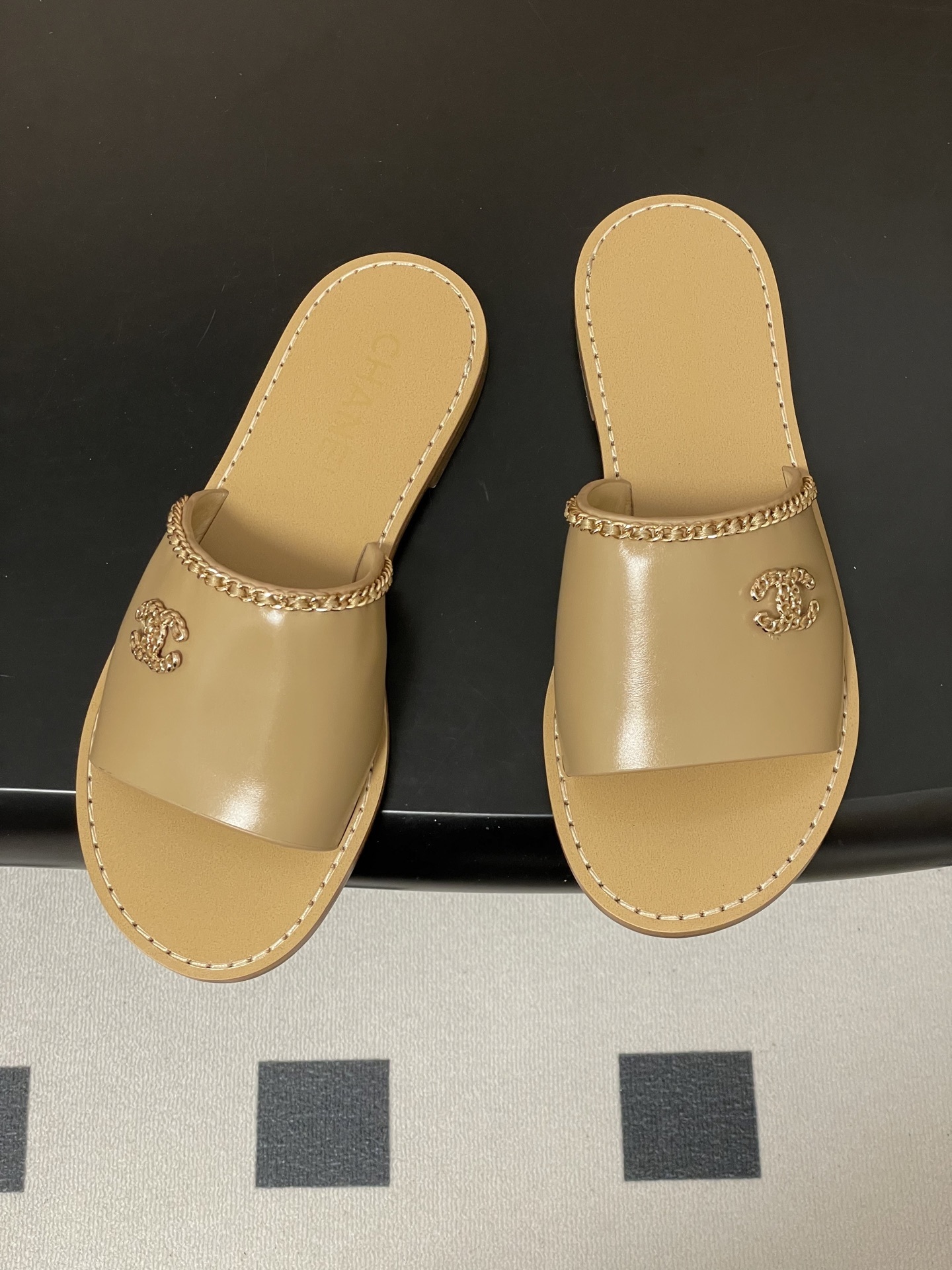 Luxury beige Chanel leather flat sandals with gold chain trim and interlocking CC logo on a black surface. - Hình 2