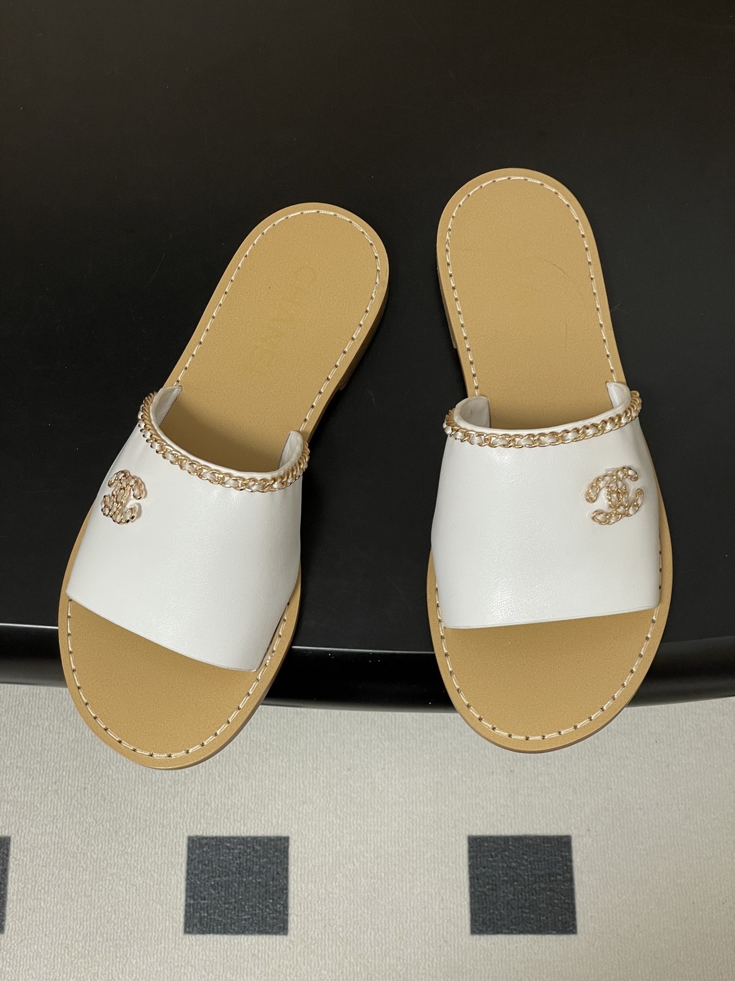 Pair of Chanel white leather flat sandals with gold chain detail and crystal embellished CC logo on a black surface. - Hình 2