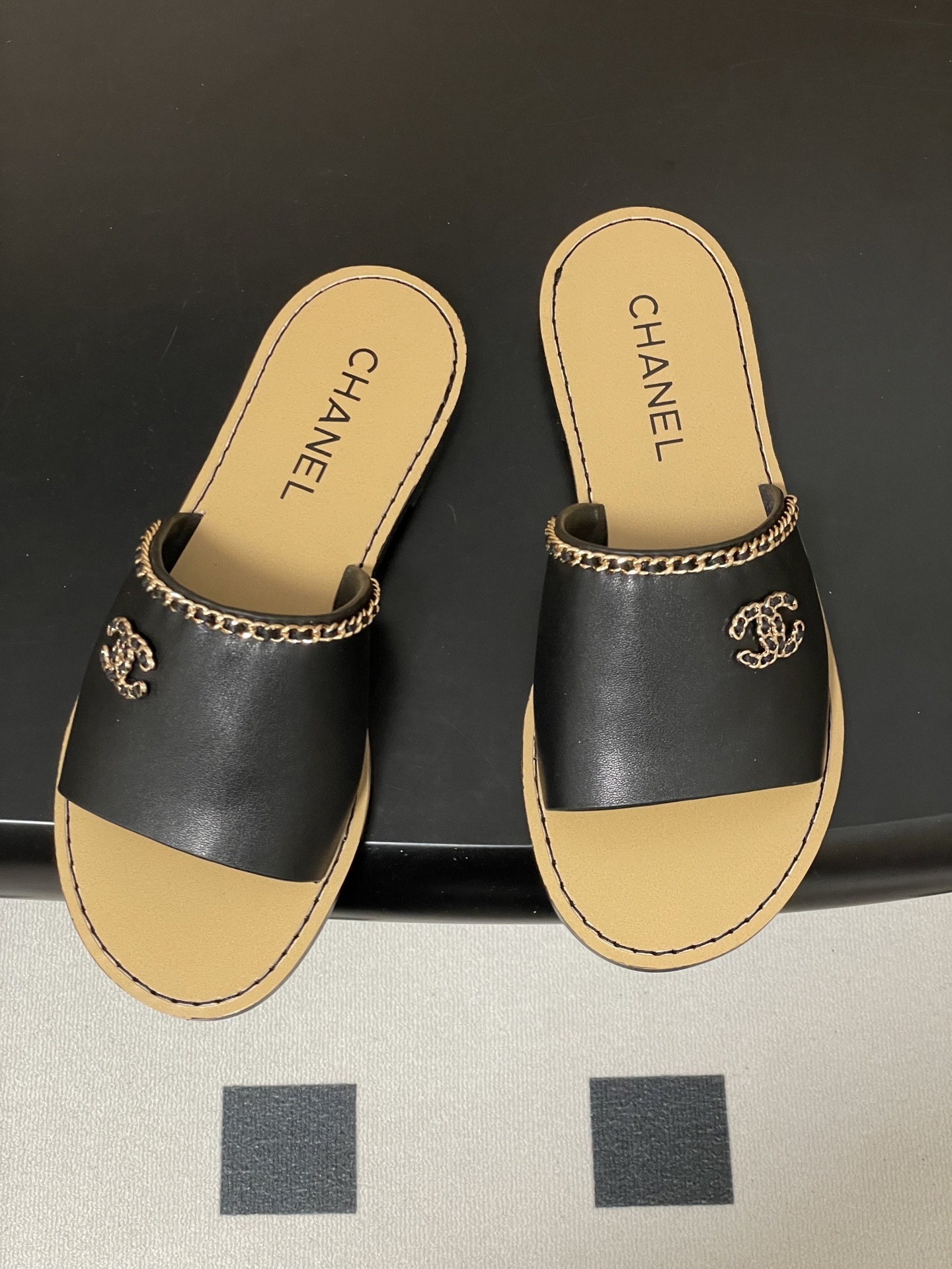 Pair of Chanel black leather flat sandals with gold chain trim and interlocking CC logo on a black surface - Hình 2