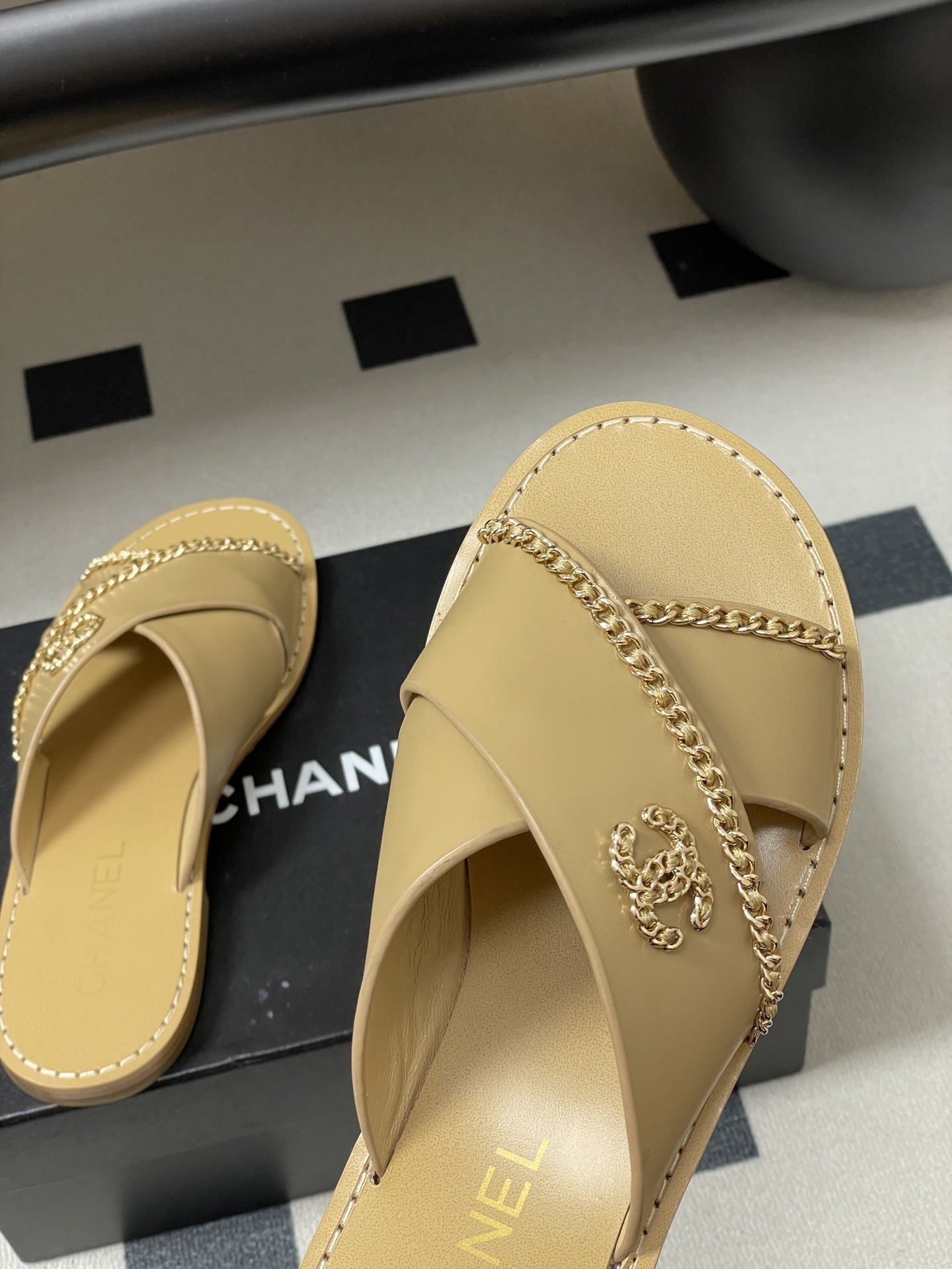 Pair of Chanel beige leather flat sandals with criss-cross straps and gold chain CC logo detailing on a black surface. - Hình 9