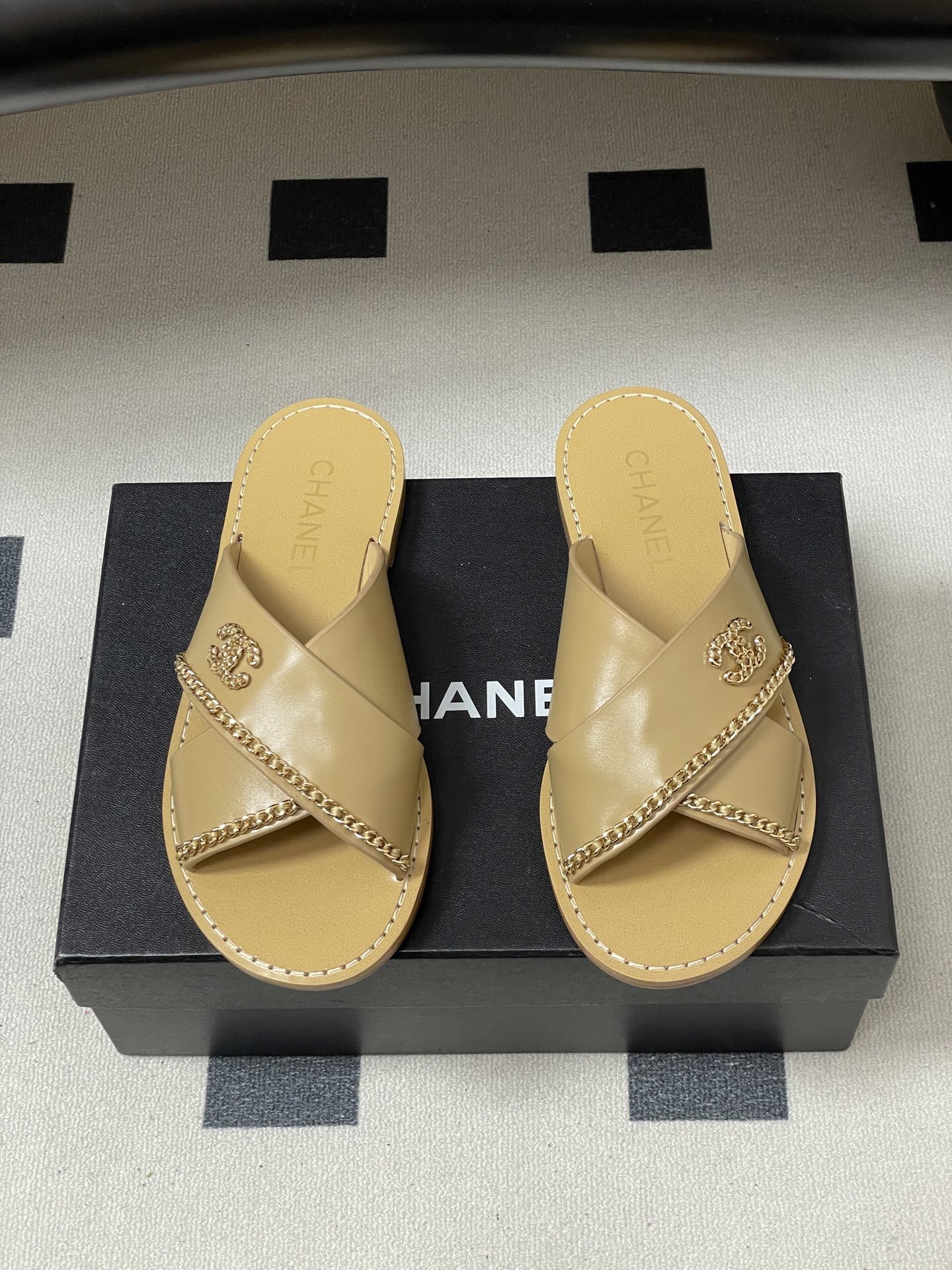 Pair of Chanel beige leather flat sandals with criss-cross straps and gold chain CC logo detailing on a black surface. - Hình 5