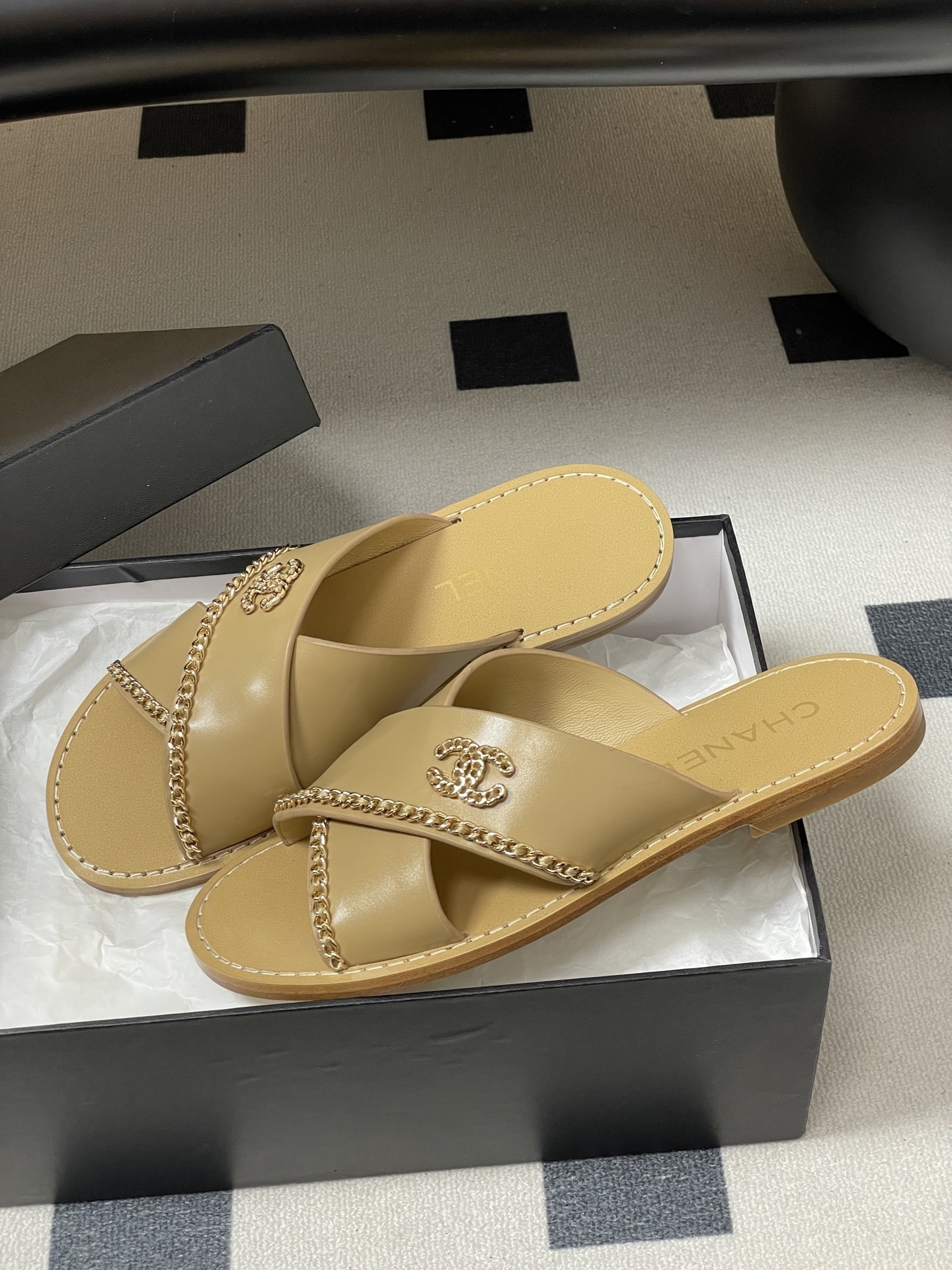 Pair of Chanel beige leather flat sandals with criss-cross straps and gold chain CC logo detailing on a black surface. - Hình 4