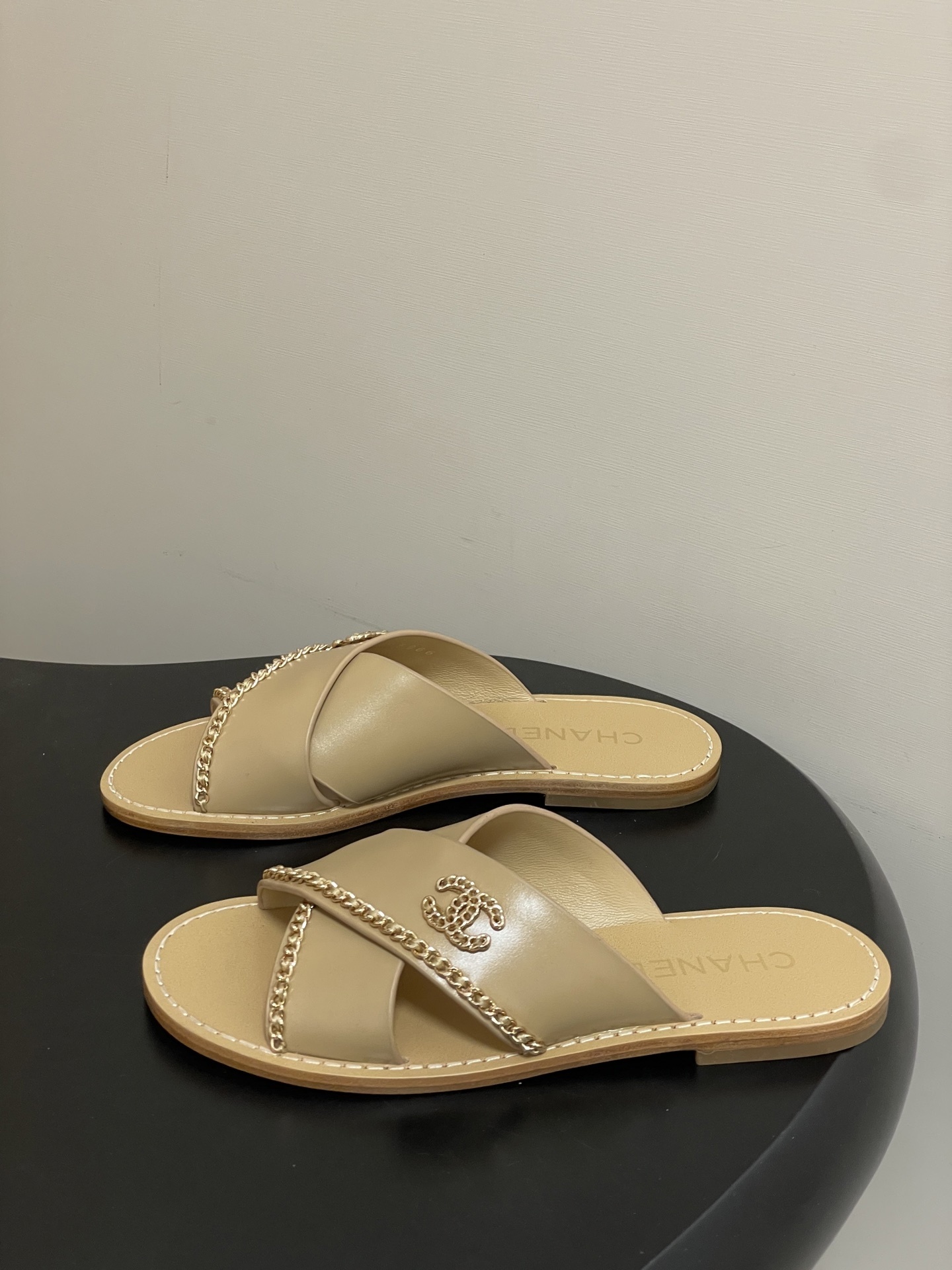 Pair of Chanel beige leather flat sandals with criss-cross straps and gold chain CC logo detailing on a black surface. - Hình 3