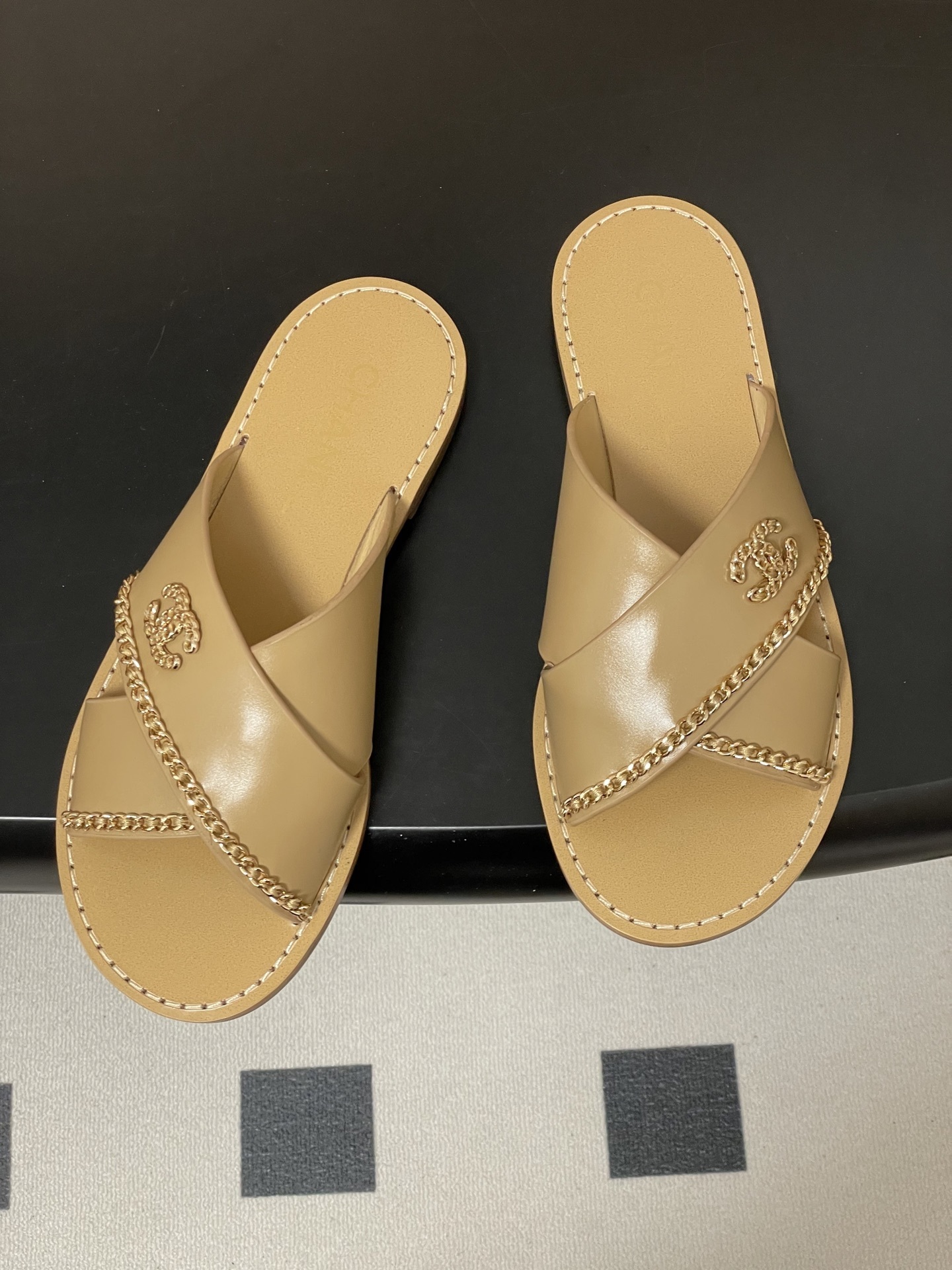 Pair of Chanel beige leather flat sandals with criss-cross straps and gold chain CC logo detailing on a black surface. - Hình 2