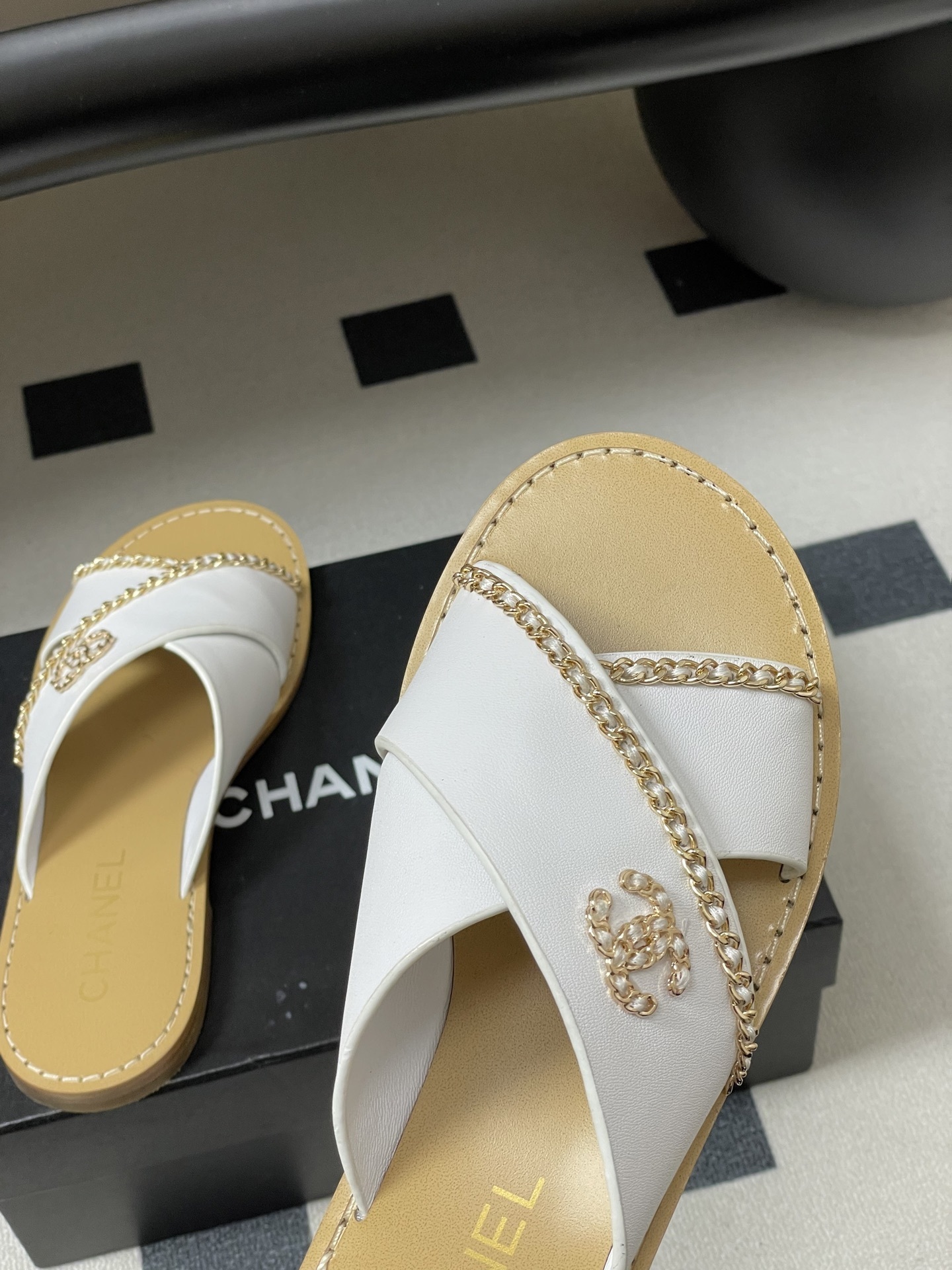 Pair of luxury Chanel white leather criss-cross sandals with gold chain detailing and logo on a black surface. - Hình 9