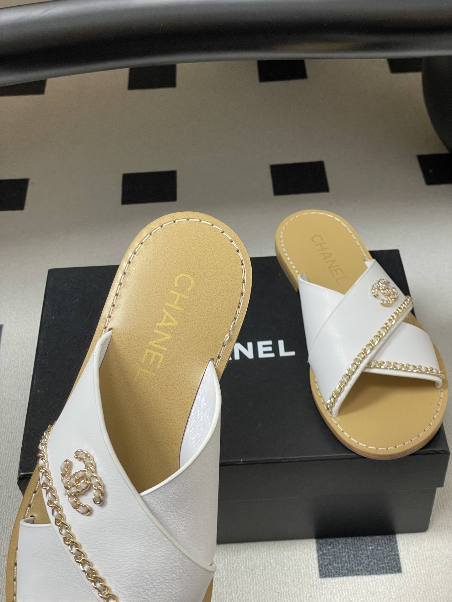 Pair of luxury Chanel white leather criss-cross sandals with gold chain detailing and logo on a black surface. - Hình 7