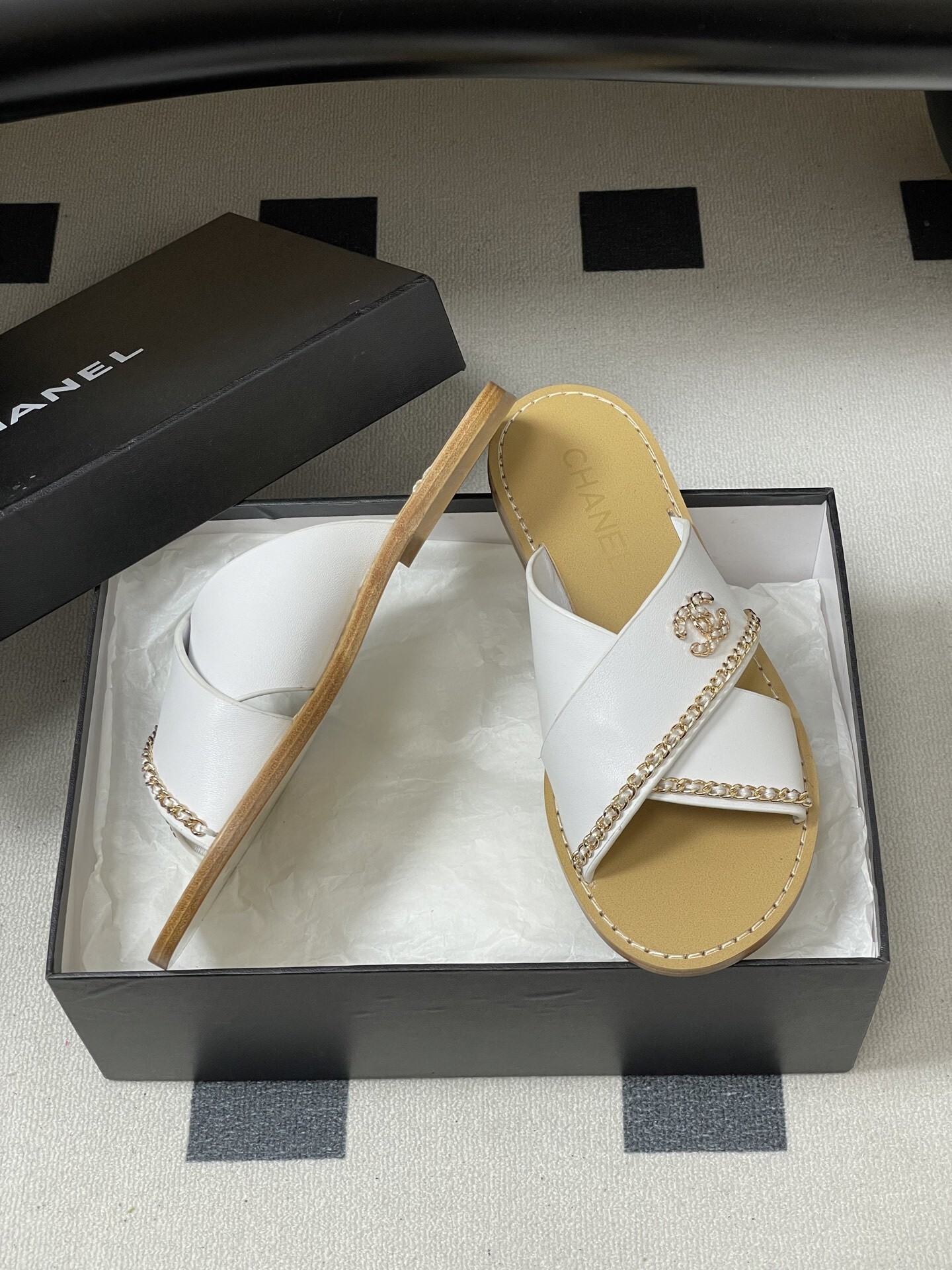 Pair of luxury Chanel white leather criss-cross sandals with gold chain detailing and logo on a black surface. - Hình 6