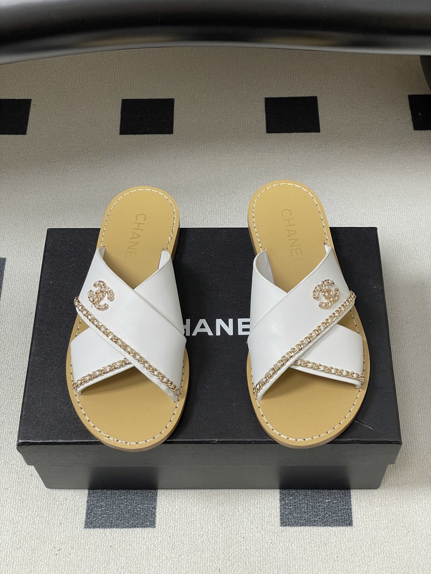 Pair of luxury Chanel white leather criss-cross sandals with gold chain detailing and logo on a black surface. - Hình 5