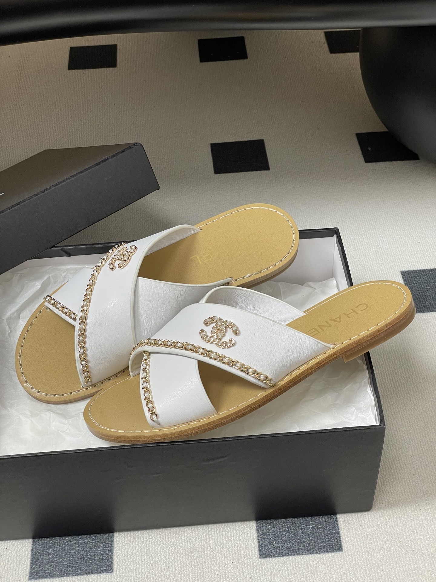 Pair of luxury Chanel white leather criss-cross sandals with gold chain detailing and logo on a black surface. - Hình 4