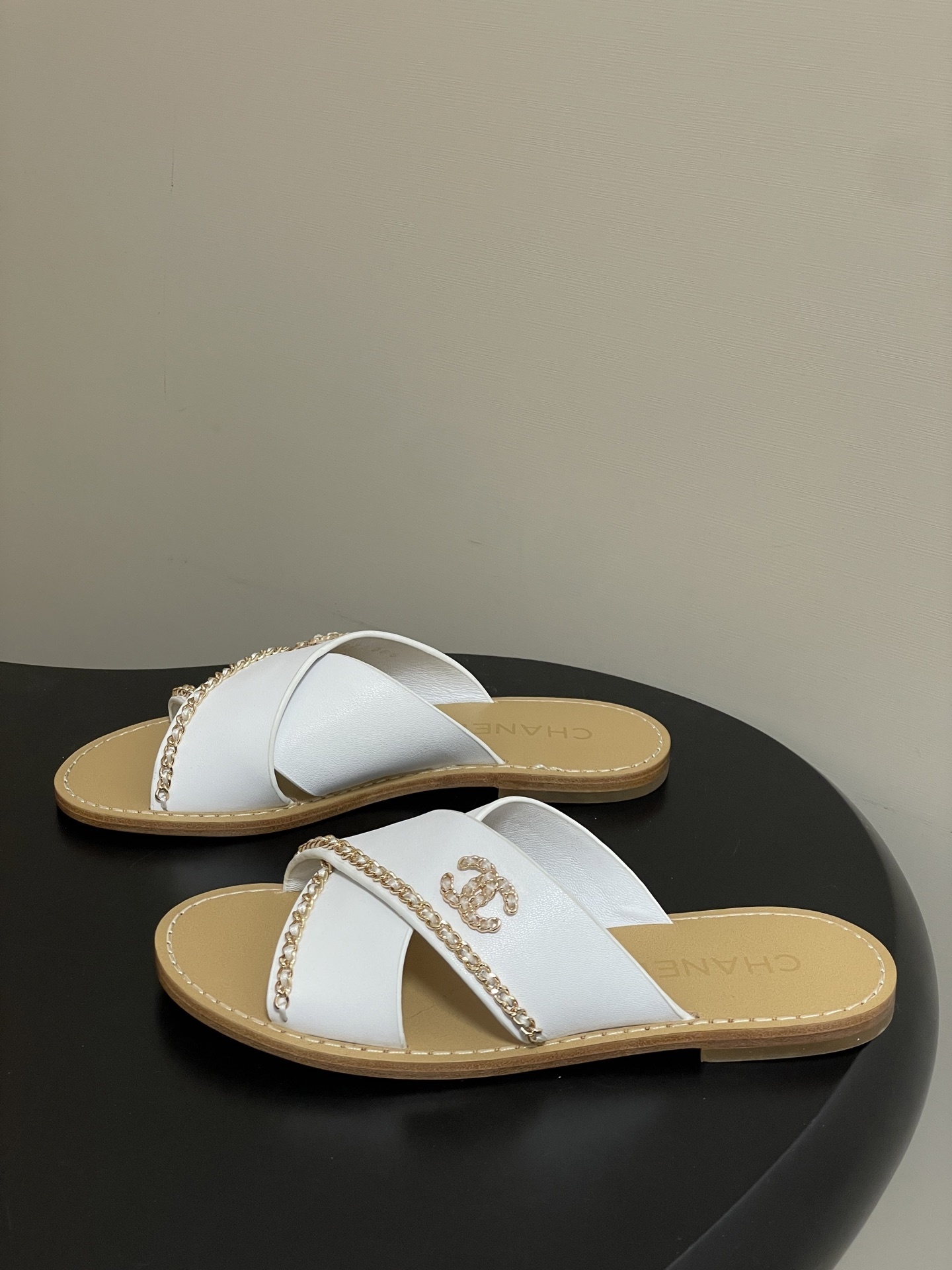 Pair of luxury Chanel white leather criss-cross sandals with gold chain detailing and logo on a black surface. - Hình 3