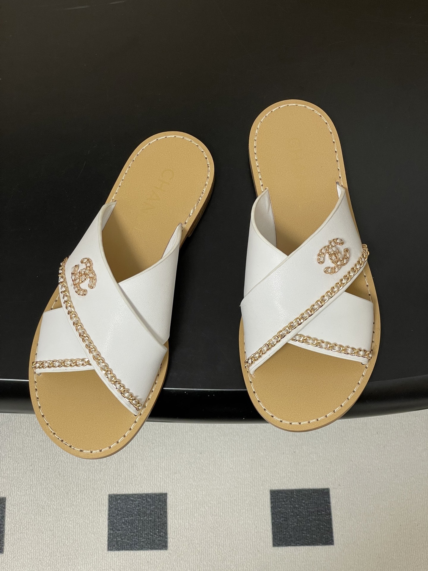 Pair of luxury Chanel white leather criss-cross sandals with gold chain detailing and logo on a black surface. - Hình 2