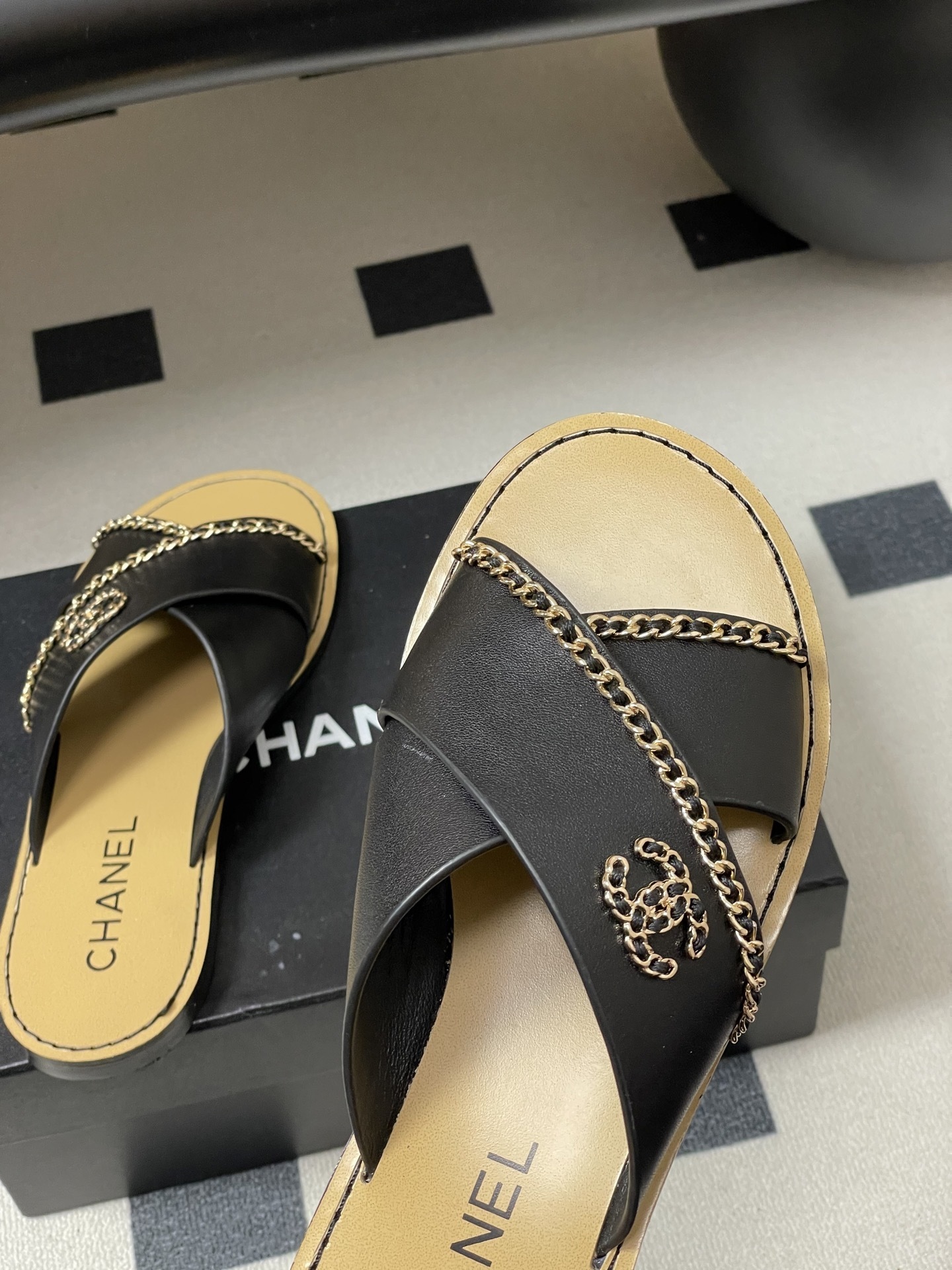 Pair of Chanel black leather criss-cross flat sandals with gold chain accents and interlocking CC logo on a black surface. - Hình 9