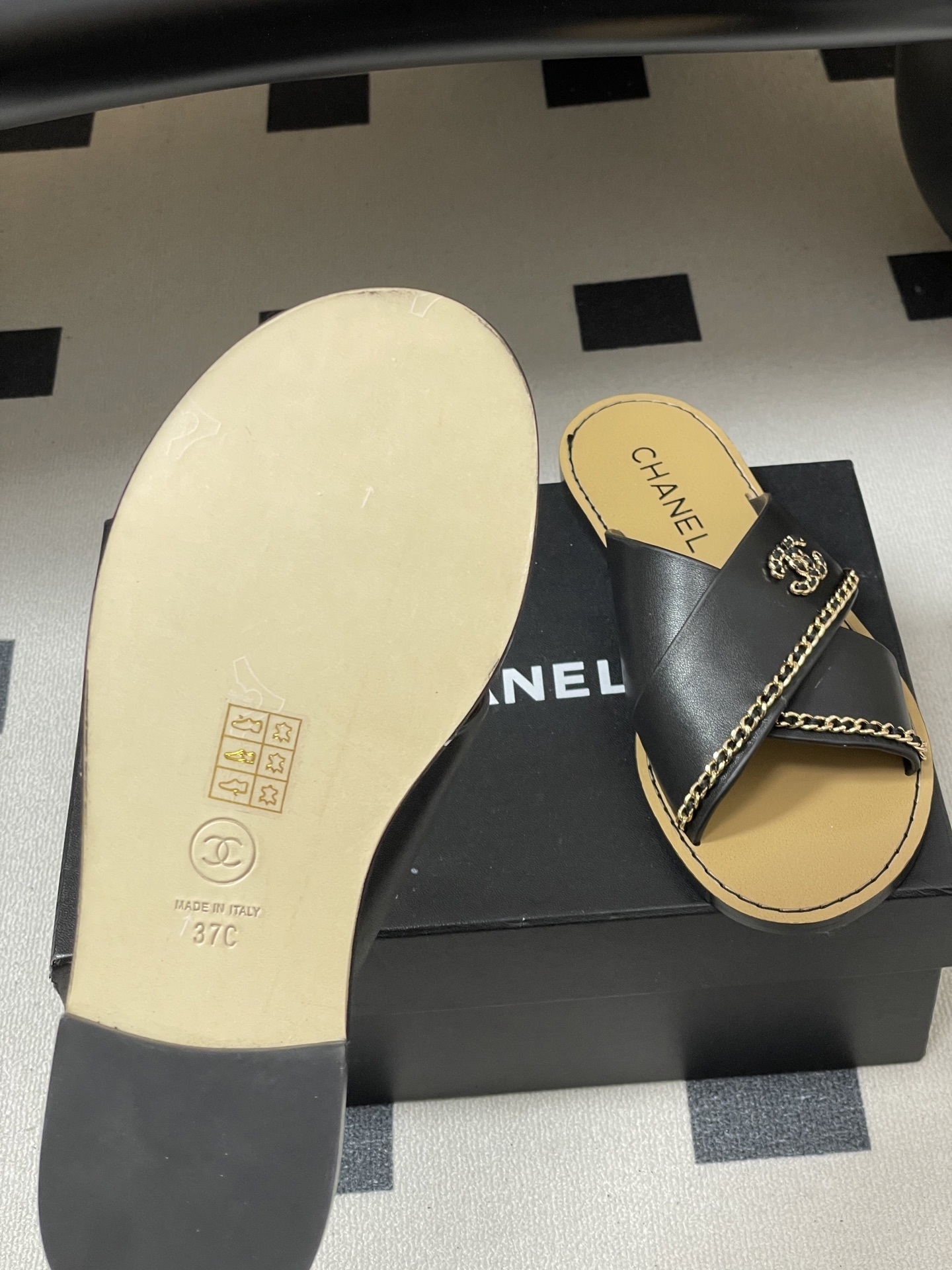 Pair of Chanel black leather criss-cross flat sandals with gold chain accents and interlocking CC logo on a black surface. - Hình 8