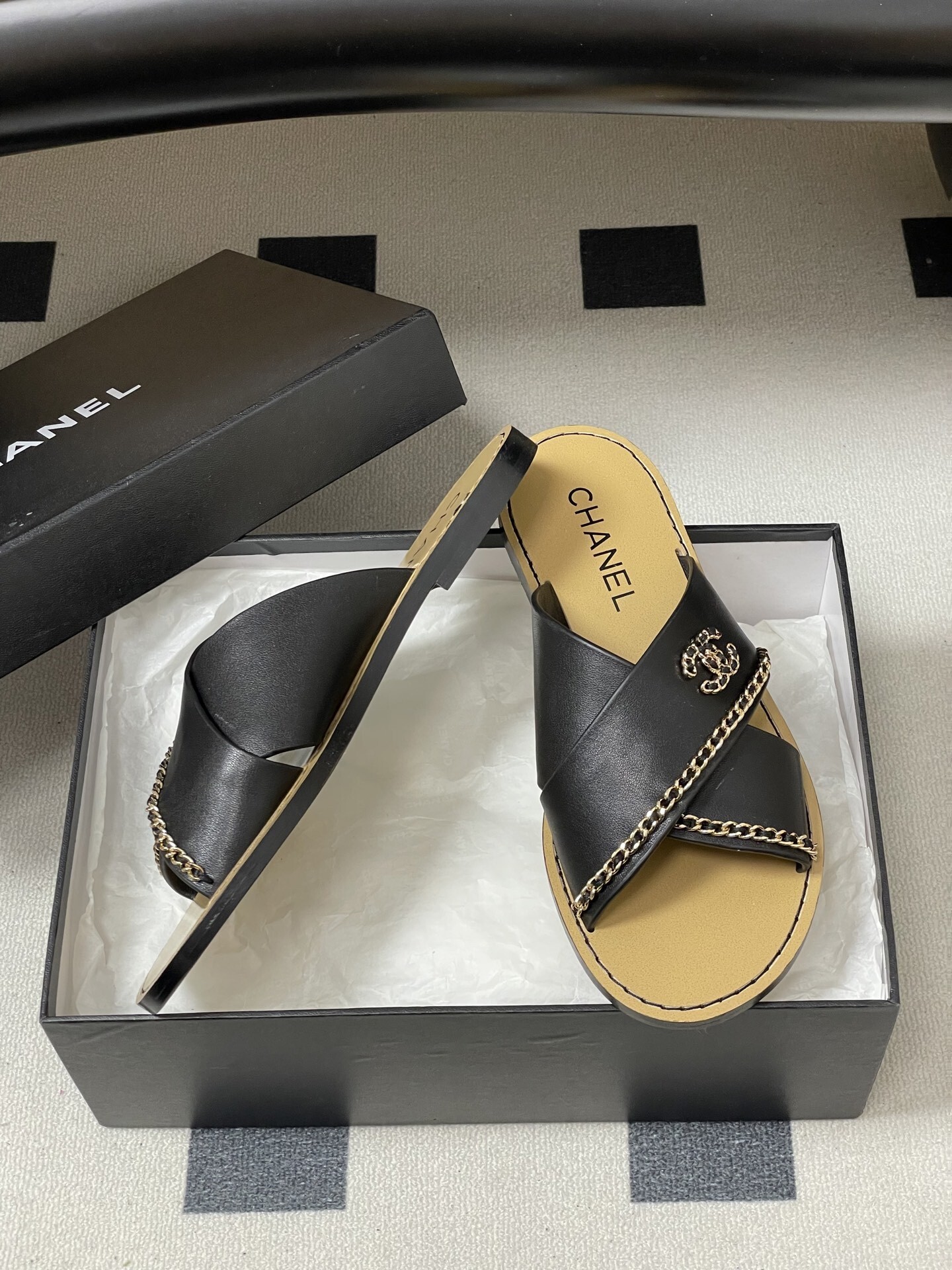 Pair of Chanel black leather criss-cross flat sandals with gold chain accents and interlocking CC logo on a black surface. - Hình 6