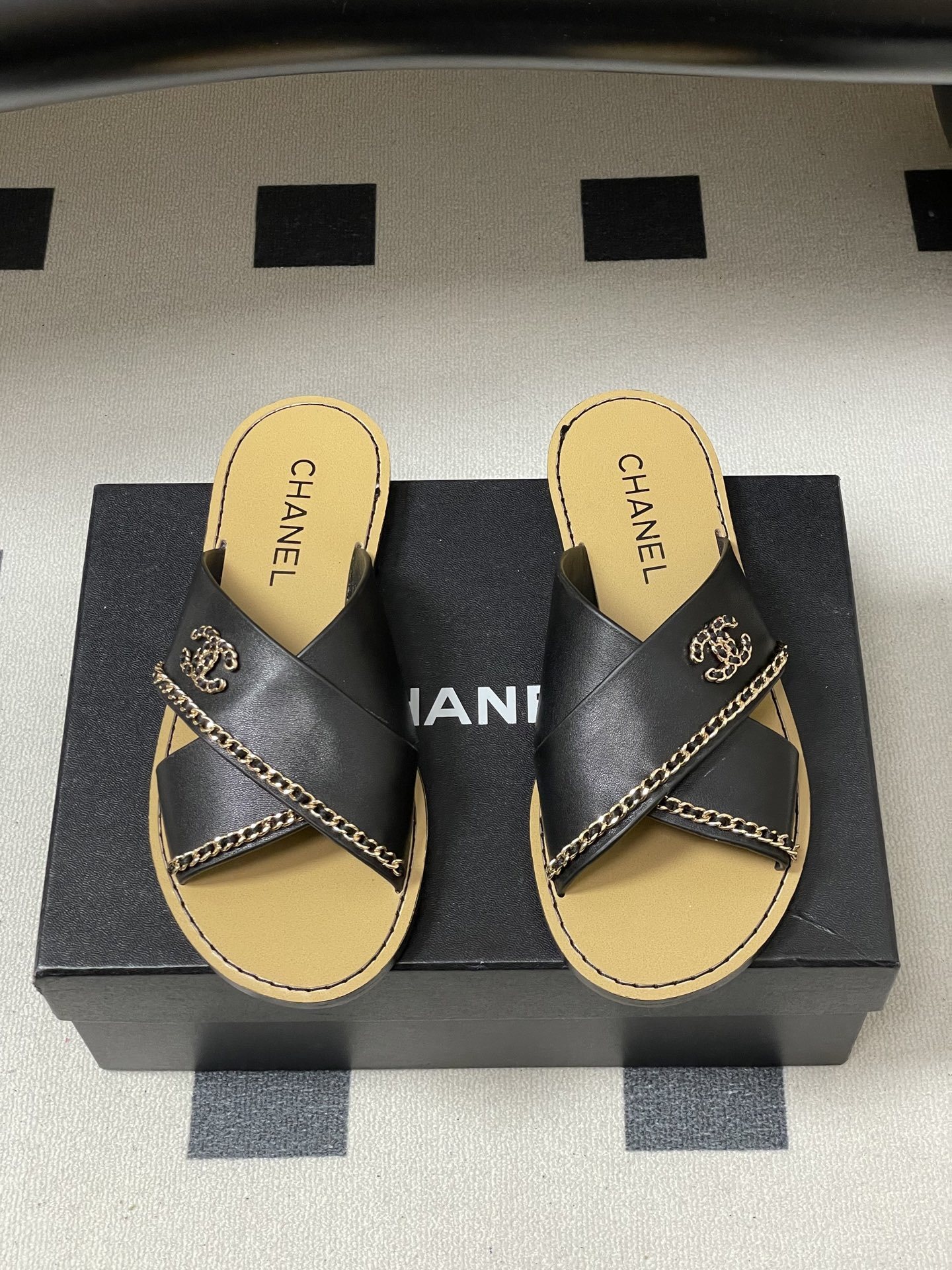 Pair of Chanel black leather criss-cross flat sandals with gold chain accents and interlocking CC logo on a black surface. - Hình 5