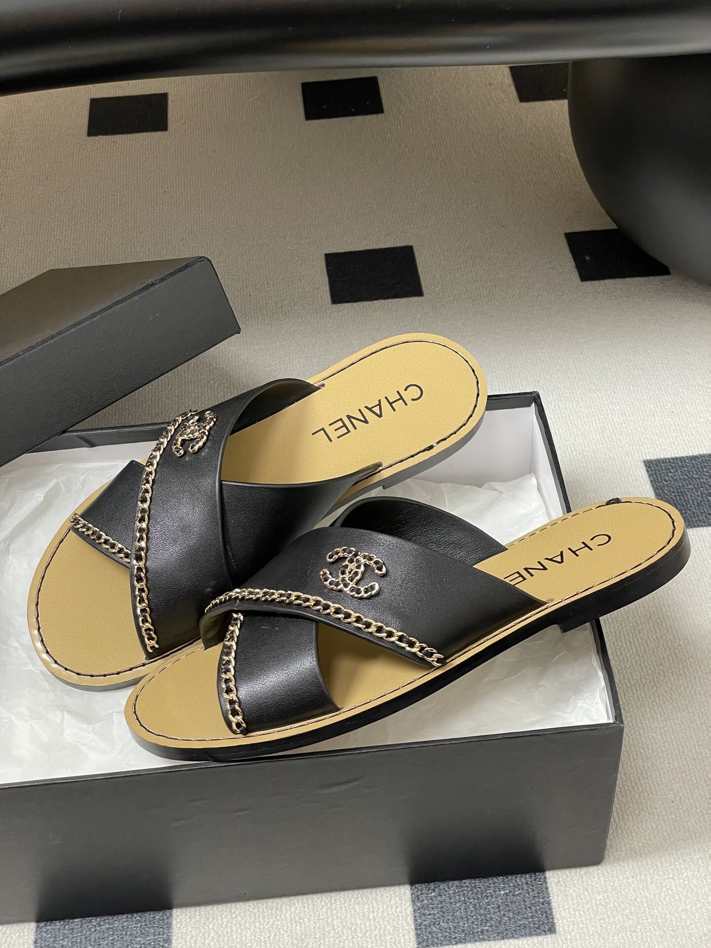 Pair of Chanel black leather criss-cross flat sandals with gold chain accents and interlocking CC logo on a black surface. - Hình 4