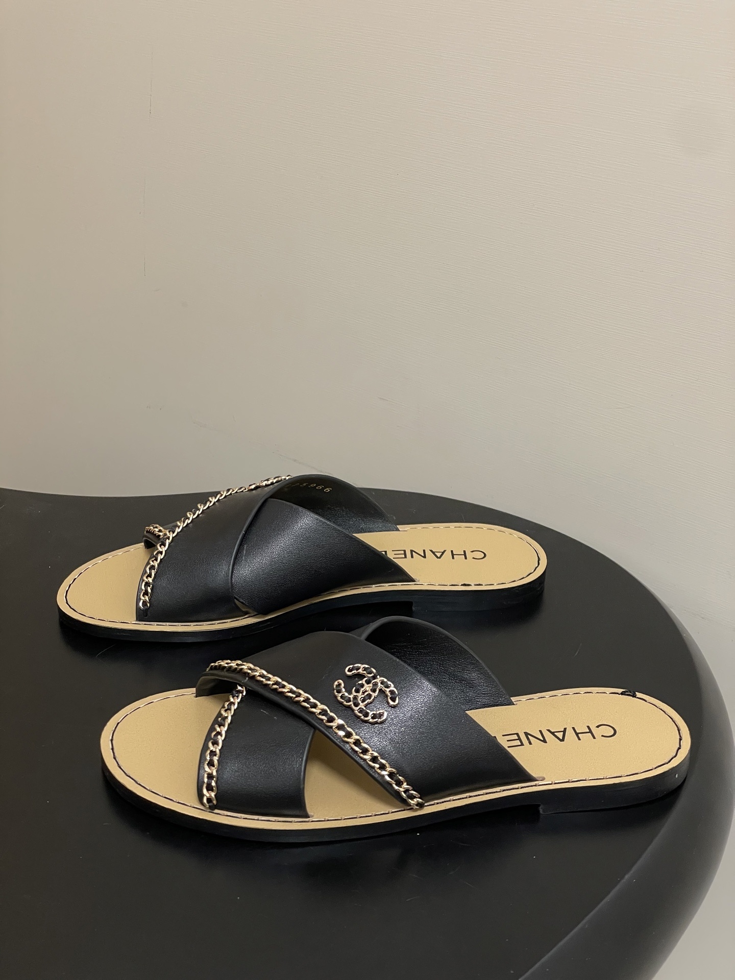 Pair of Chanel black leather criss-cross flat sandals with gold chain accents and interlocking CC logo on a black surface. - Hình 3