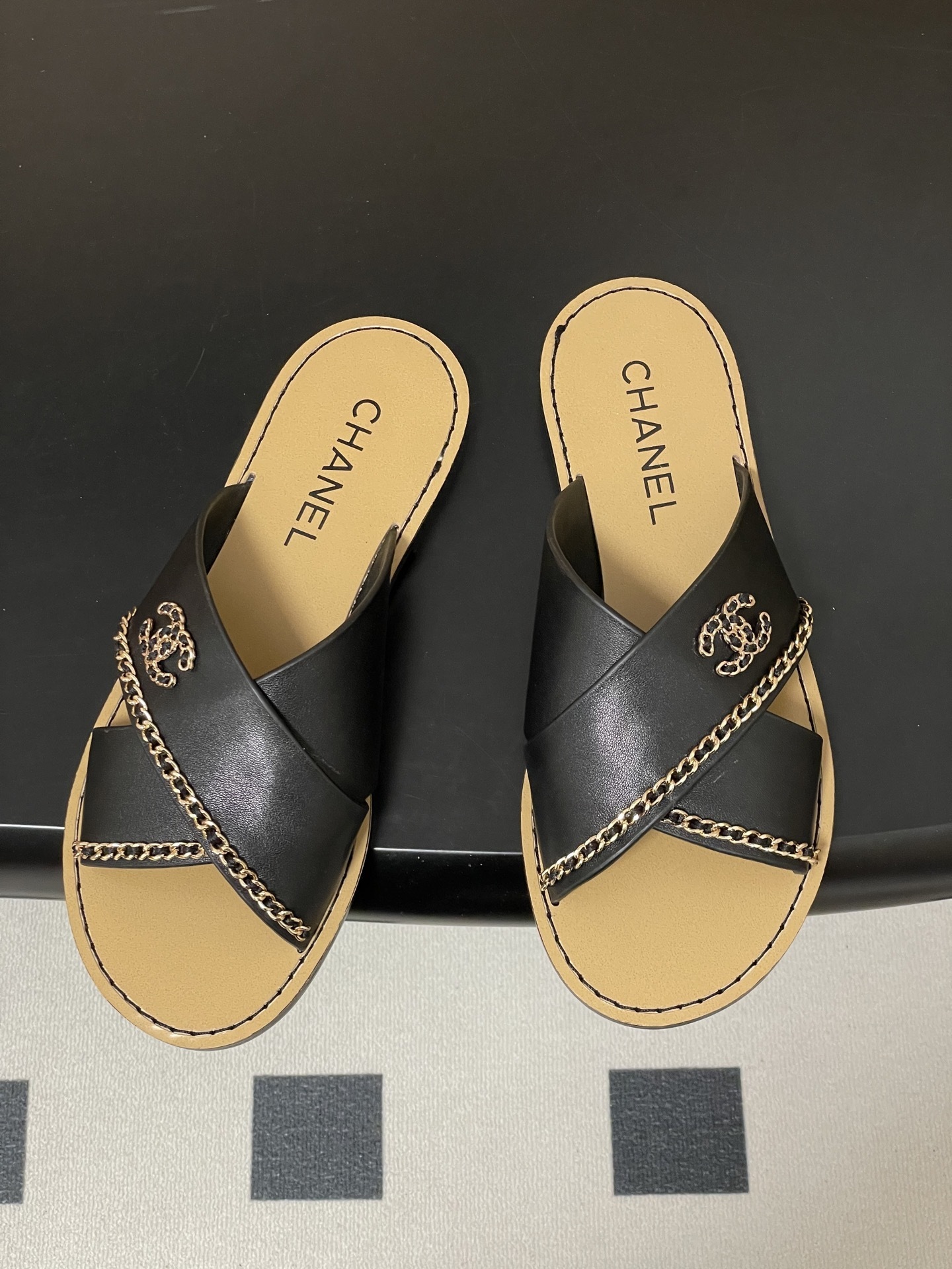 Pair of Chanel black leather criss-cross flat sandals with gold chain accents and interlocking CC logo on a black surface. - Hình 2