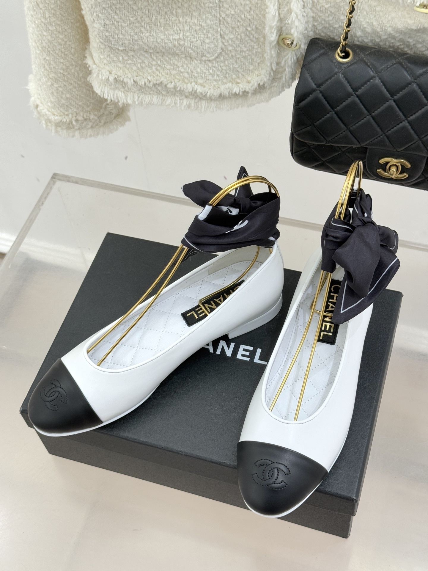 Luxury white Chanel ballerina flats with black toe caps and black silk ribbon bows displayed on a black brand box. - Hình 2