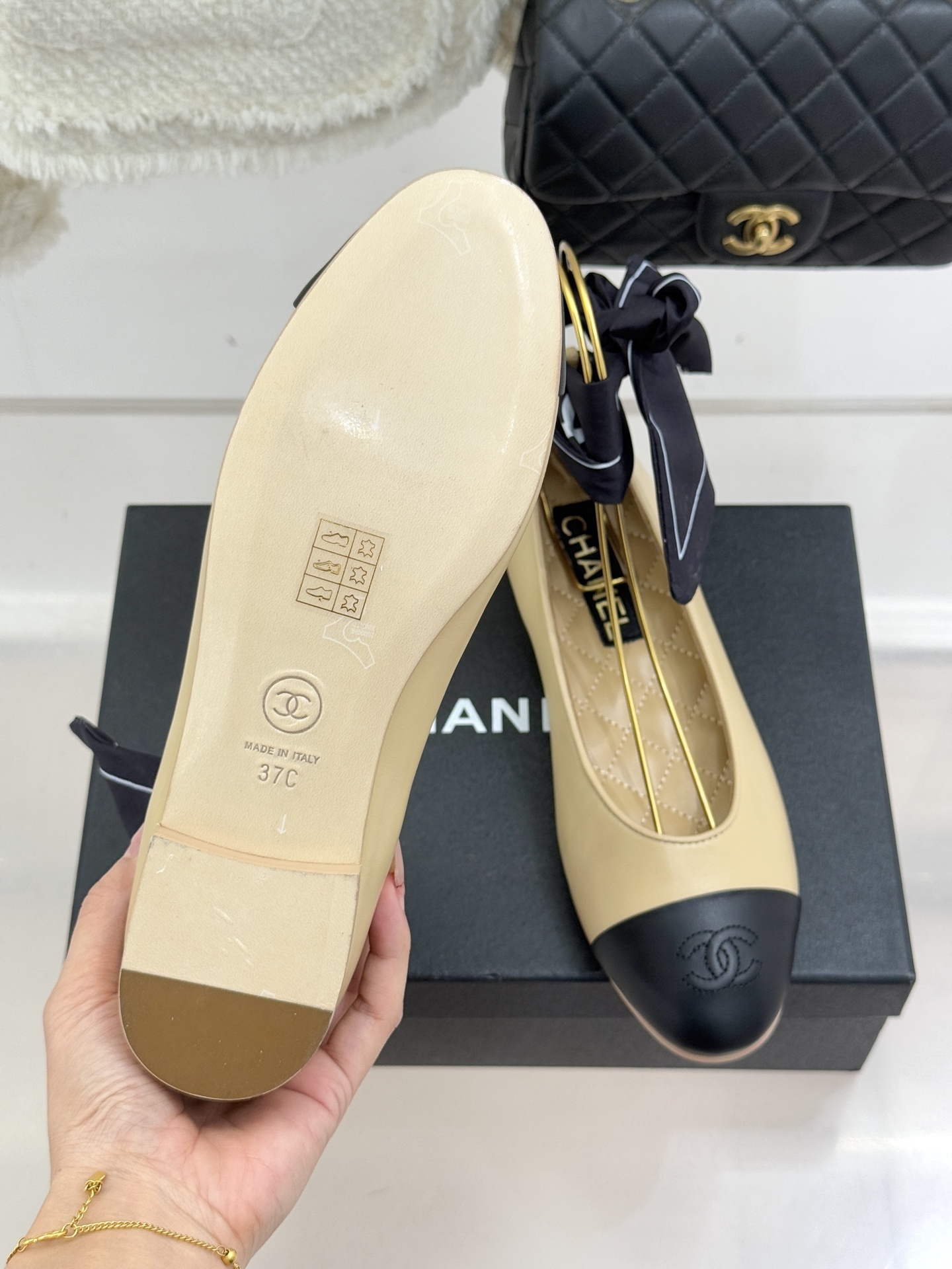 Pair of beige and black leather ballet flats with black silk ribbons on a black box next to a white tweed jacket and quilted bag - Hình 9