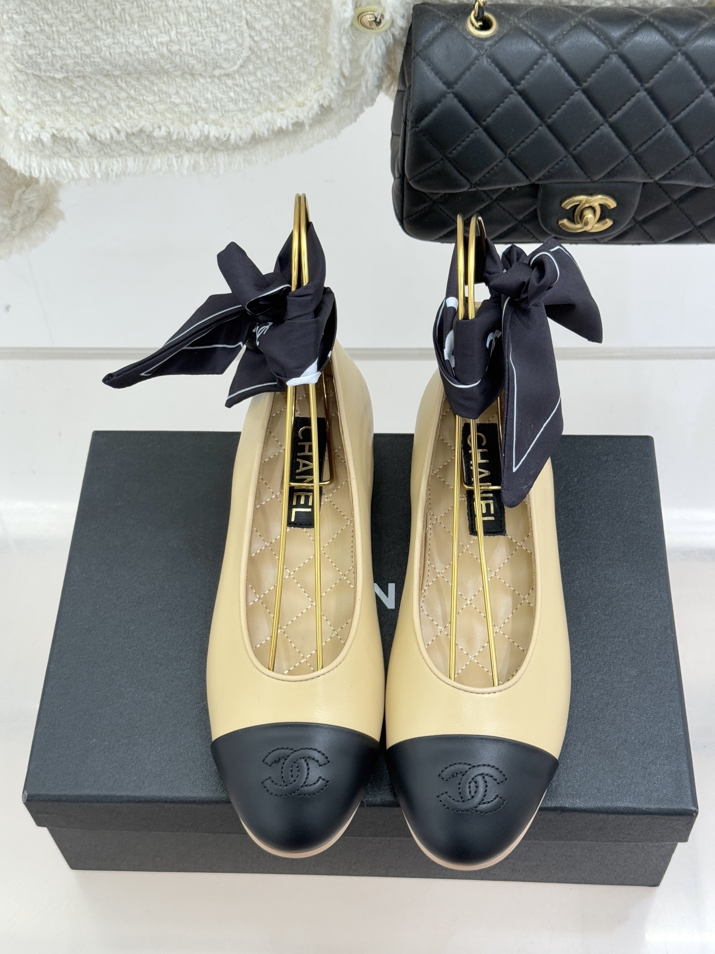 Pair of beige and black leather ballet flats with black silk ribbons on a black box next to a white tweed jacket and quilted bag - Hình 4