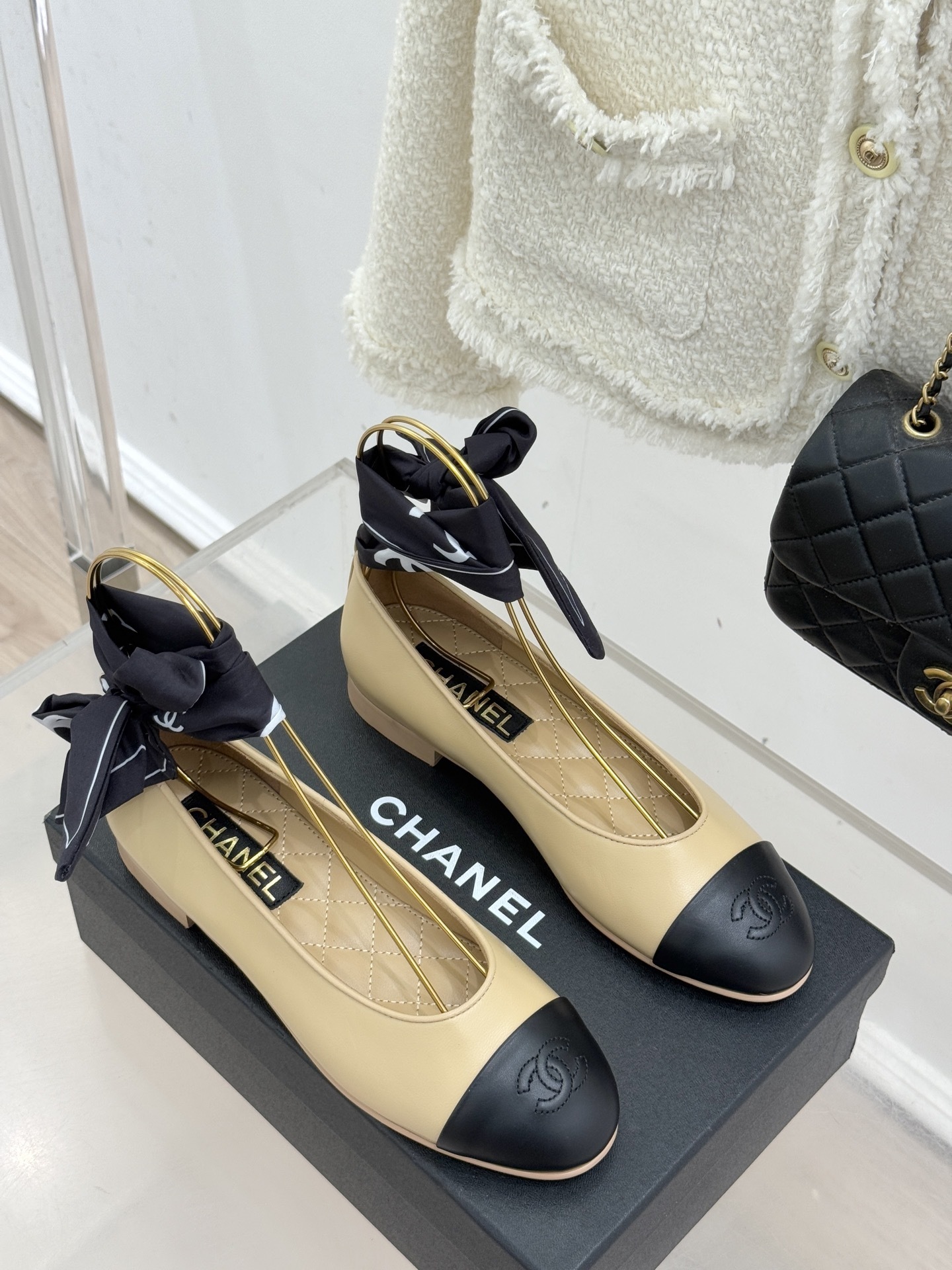 Pair of beige and black leather ballet flats with black silk ribbons on a black box next to a white tweed jacket and quilted bag - Hình 3