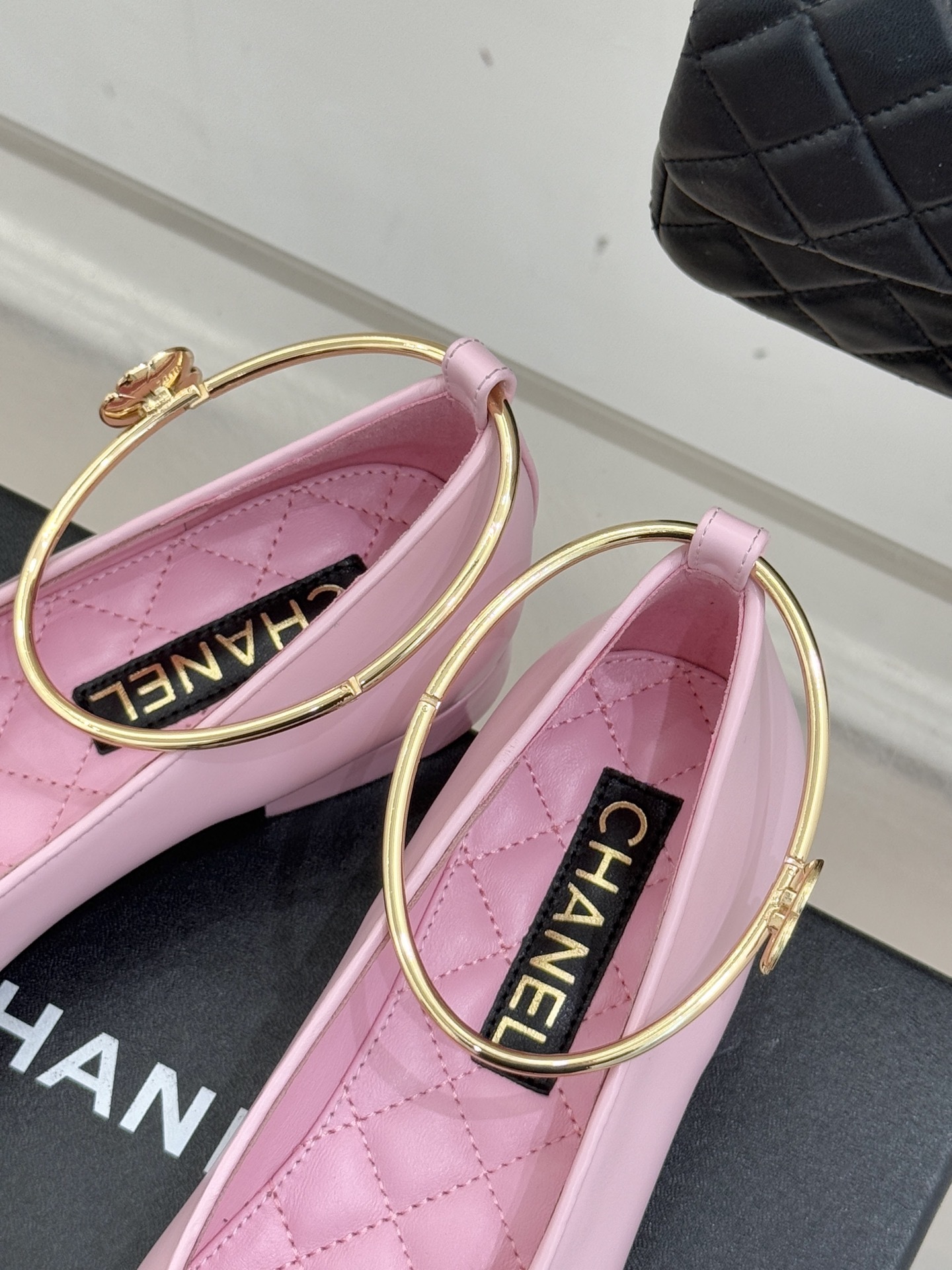 A pair of Chanel pink leather ballerina flats with black cap-toes and gold-tone metal ankle rings resting on a black box. - Hình 8