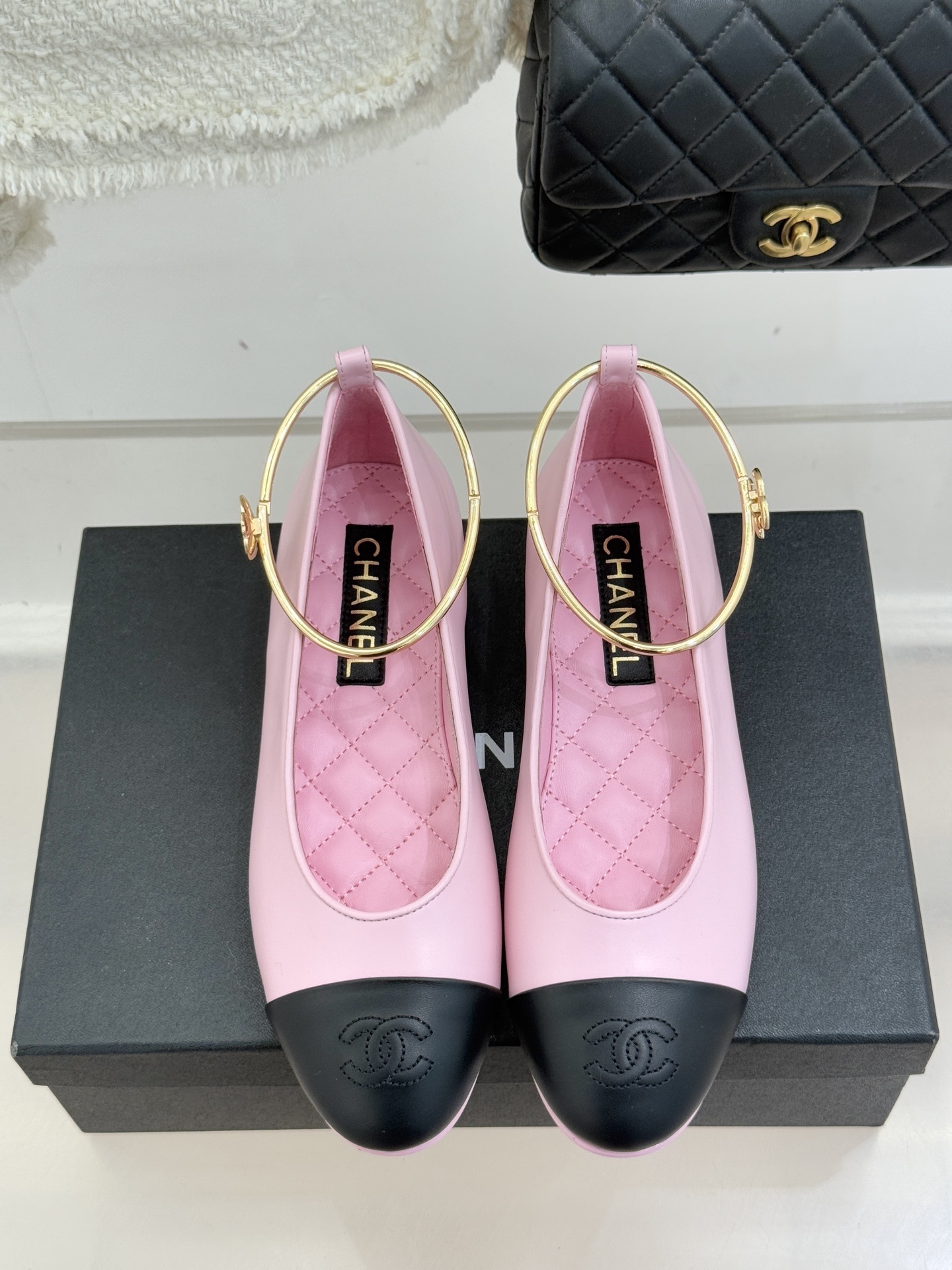 A pair of Chanel pink leather ballerina flats with black cap-toes and gold-tone metal ankle rings resting on a black box. - Hình 4