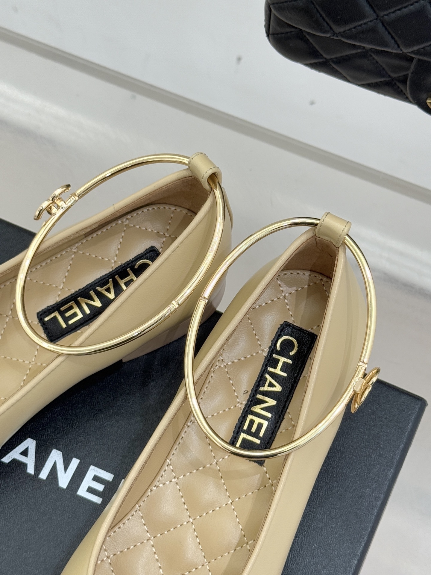 Pair of luxury Chanel two-tone beige and black ballet flats with gold circular ankle straps on a black shoe box. - Hình 8