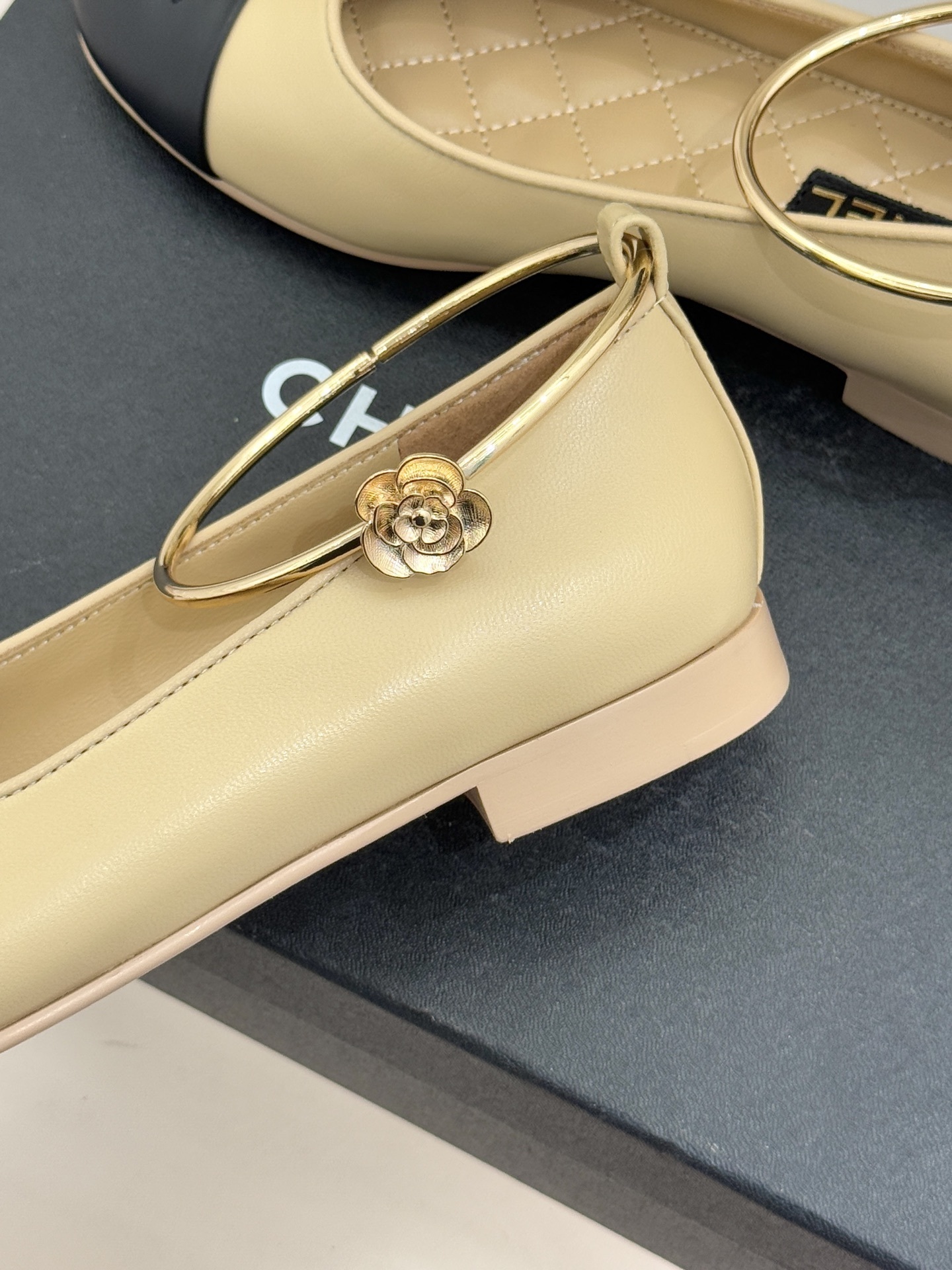 Pair of luxury Chanel two-tone beige and black ballet flats with gold circular ankle straps on a black shoe box. - Hình 6