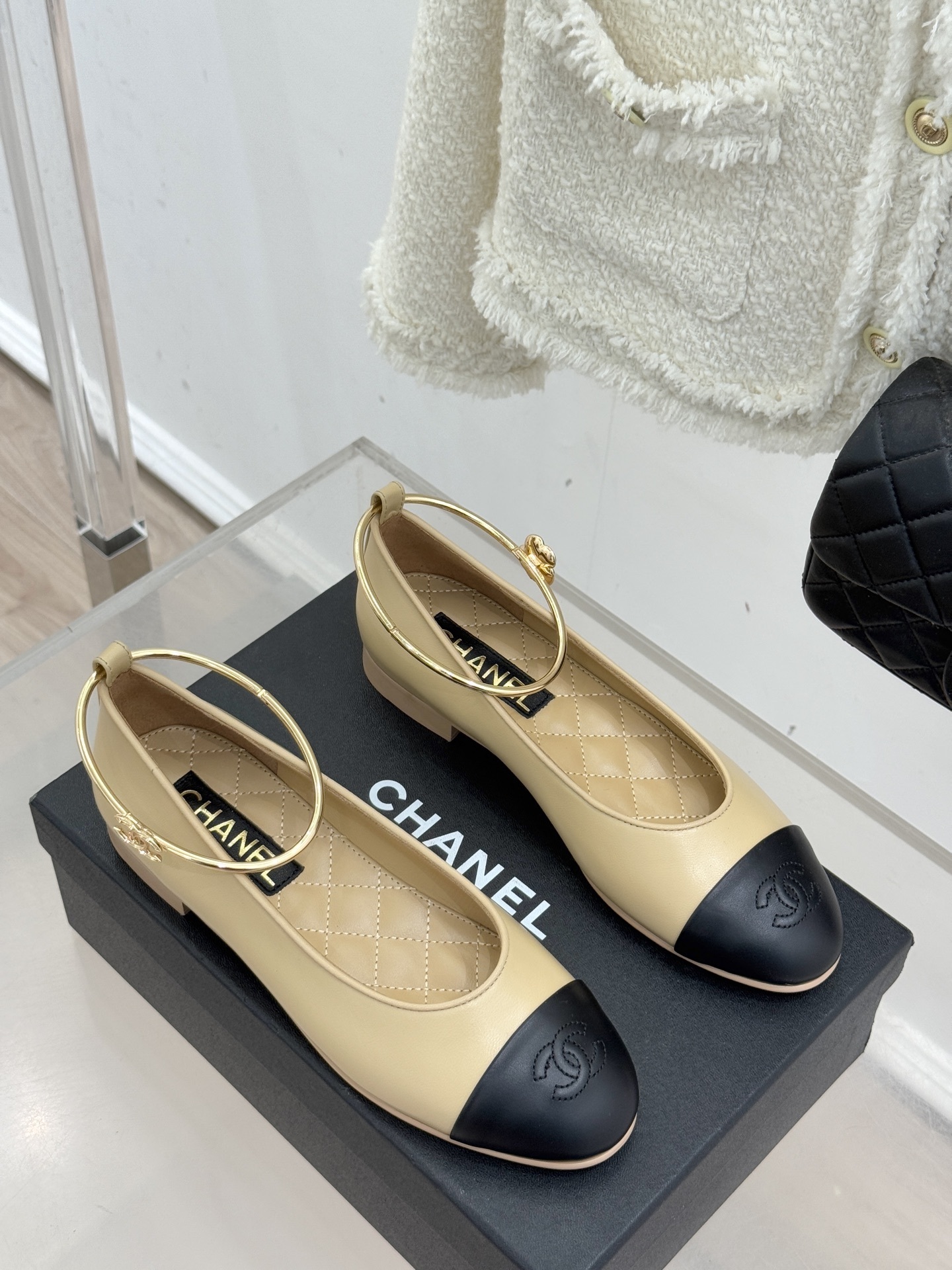 Pair of luxury Chanel two-tone beige and black ballet flats with gold circular ankle straps on a black shoe box. - Hình 3