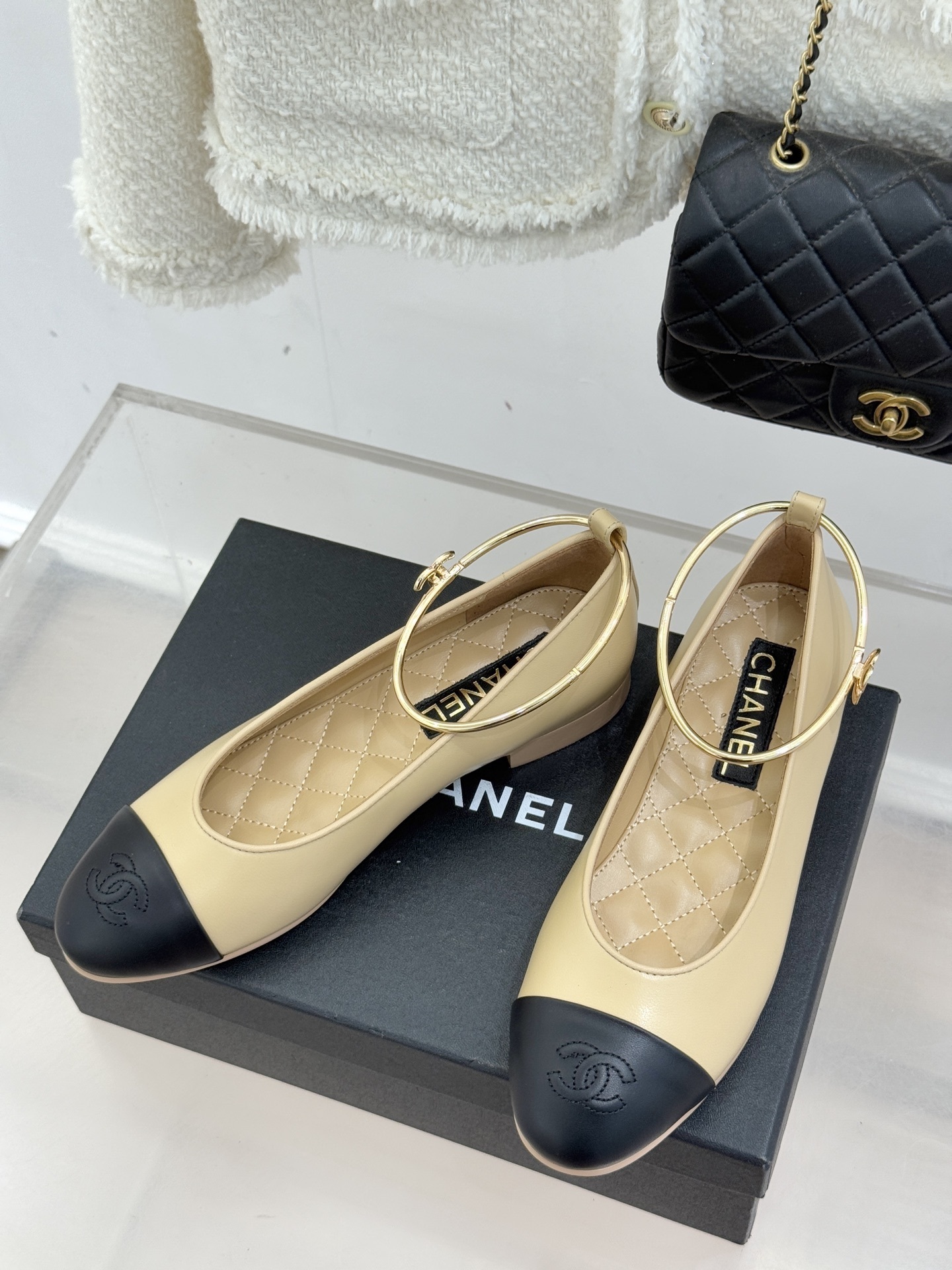 Pair of luxury Chanel two-tone beige and black ballet flats with gold circular ankle straps on a black shoe box. - Hình 2