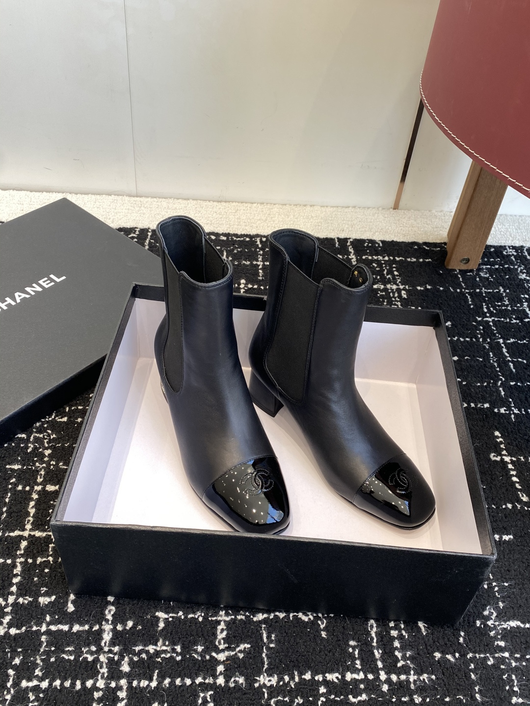 Pair of black Chanel leather ankle boots with patent leather cap-toe and CC logo on a black box. - Hình 3