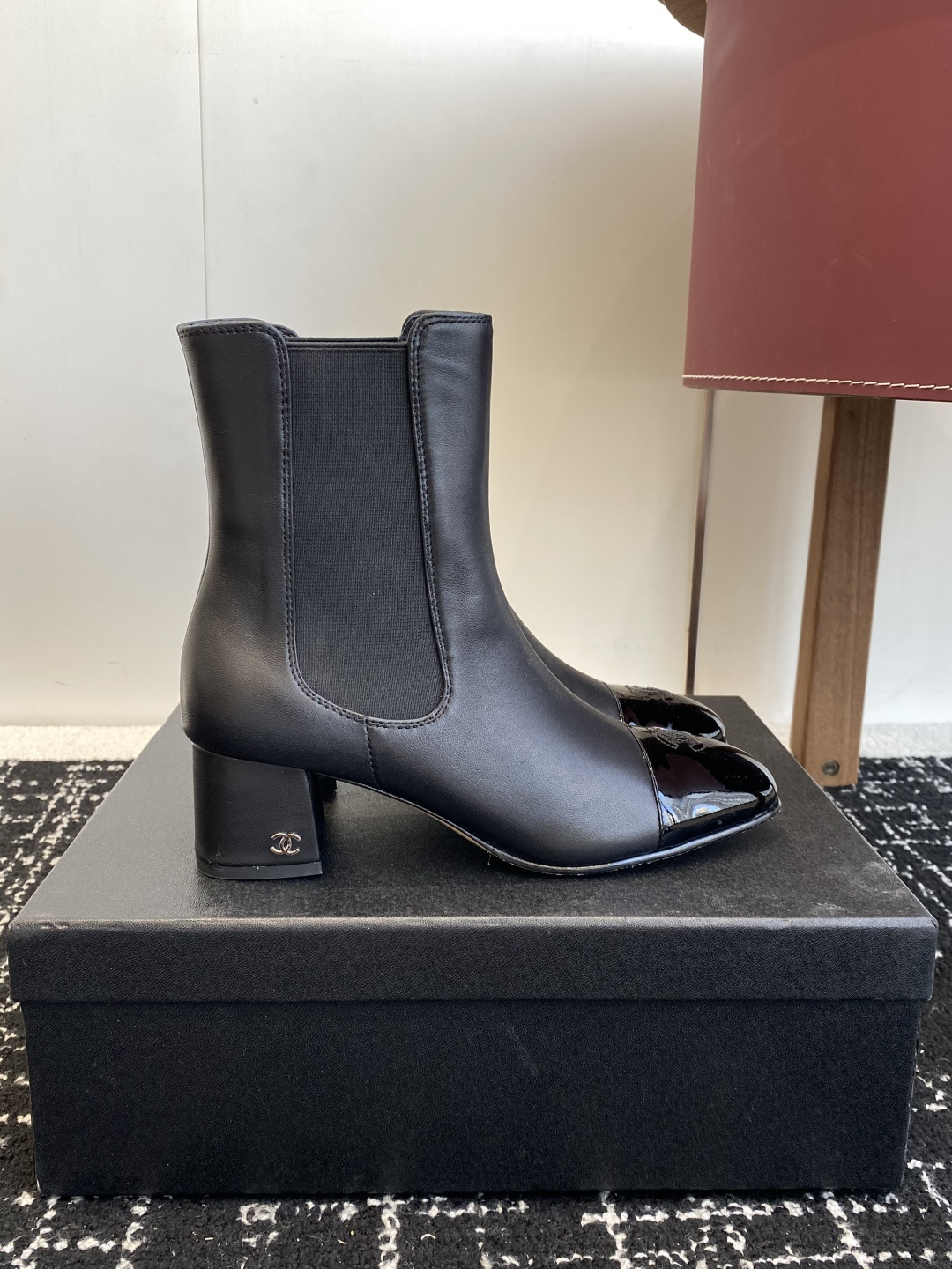 Pair of black Chanel leather ankle boots with patent leather cap-toe and CC logo on a black box. - Hình 2