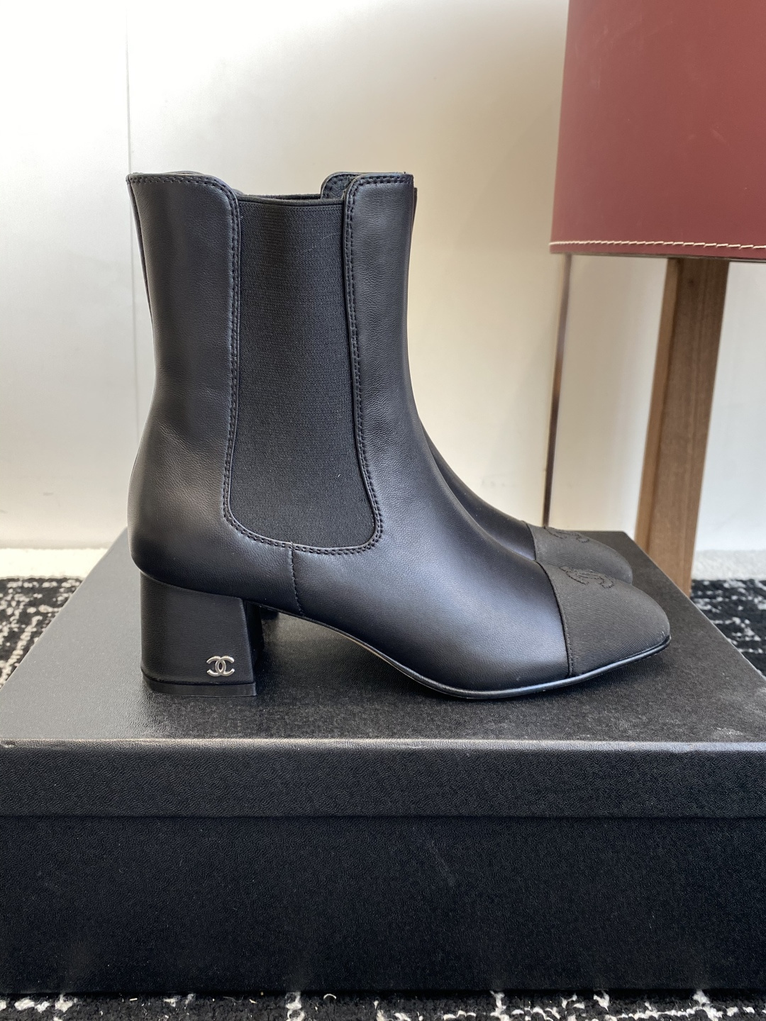 Chanel black leather Chelsea boots with signature CC logo on the toe cap and heel, displayed on a black box. - Hình 2