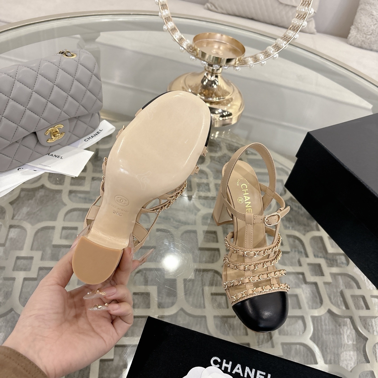Luxury Chanel beige leather block heel sandals with black cap toes and gold-tone chain straps displayed on a glass table. - Hình 9