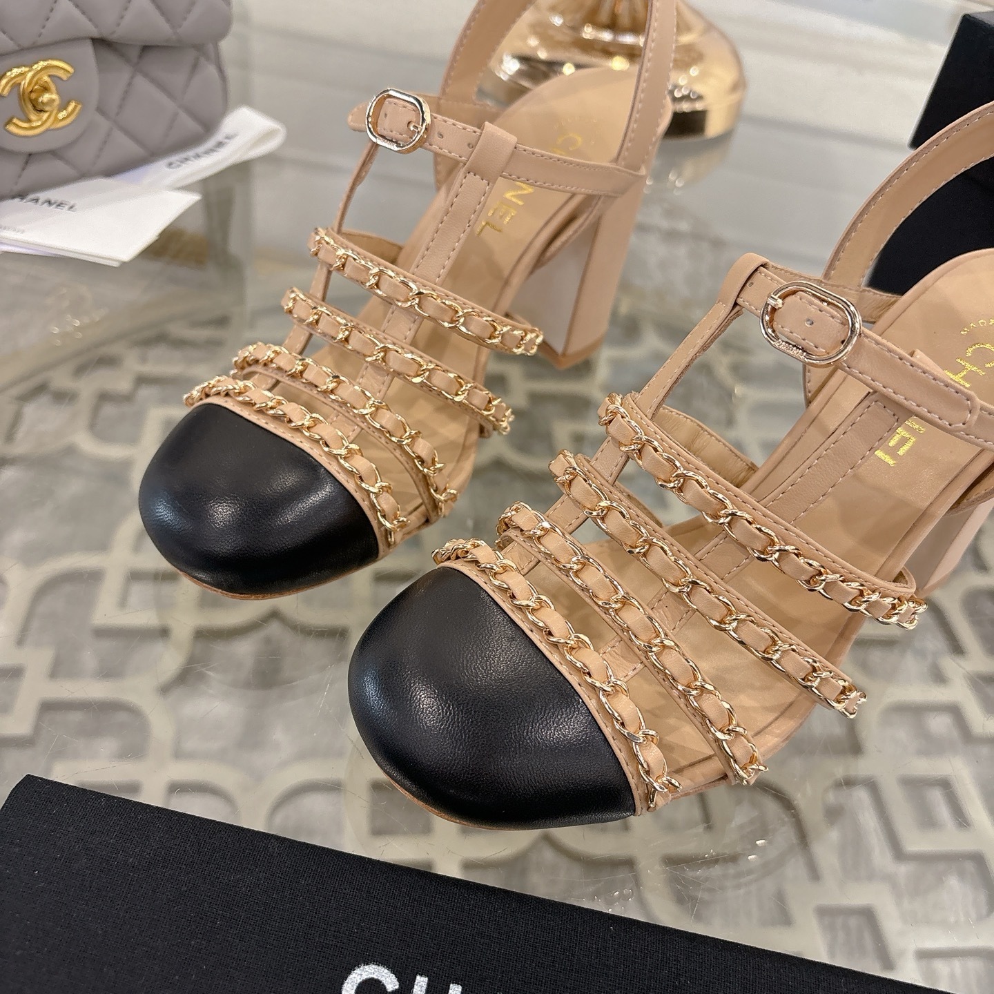 Luxury Chanel beige leather block heel sandals with black cap toes and gold-tone chain straps displayed on a glass table. - Hình 7