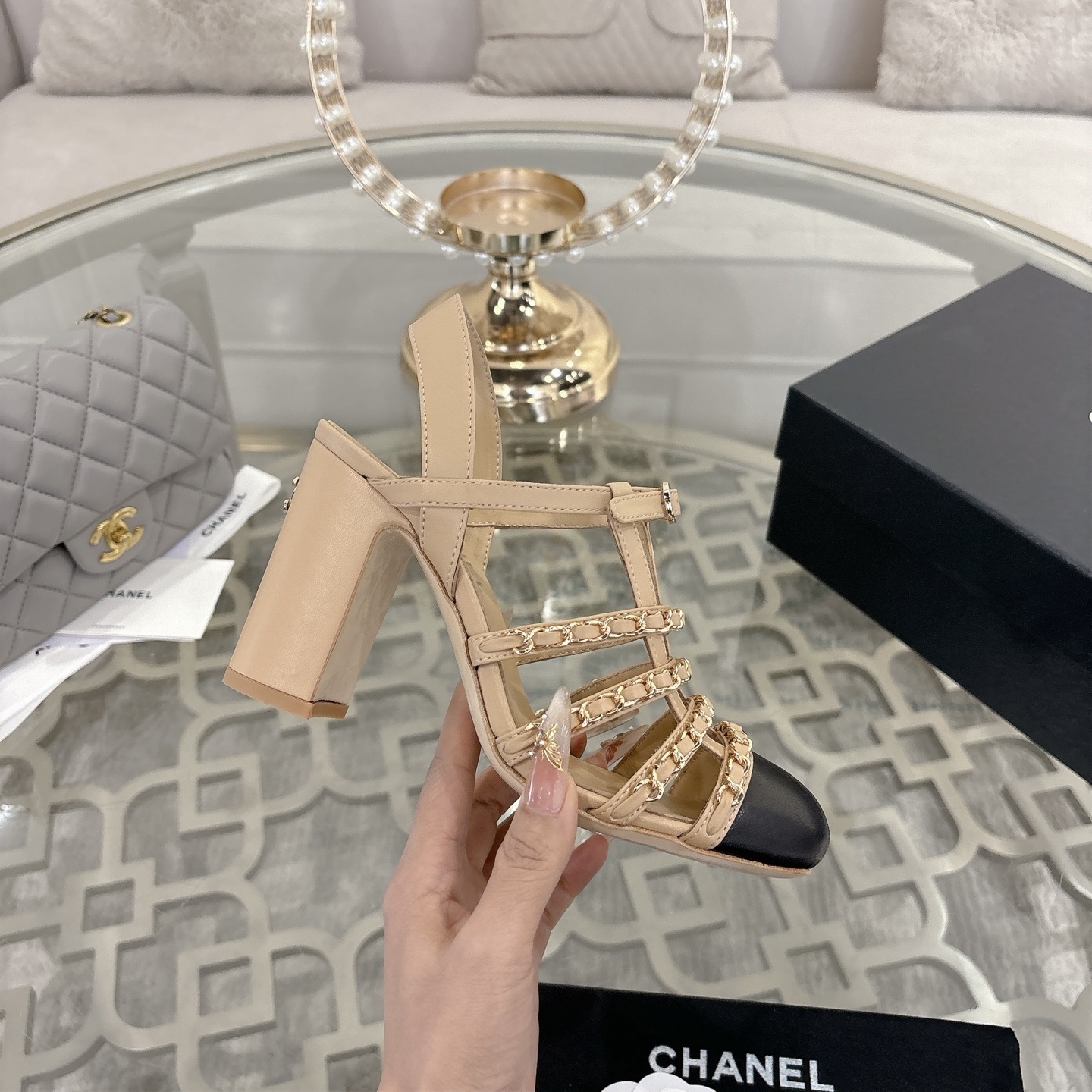 Luxury Chanel beige leather block heel sandals with black cap toes and gold-tone chain straps displayed on a glass table. - Hình 5