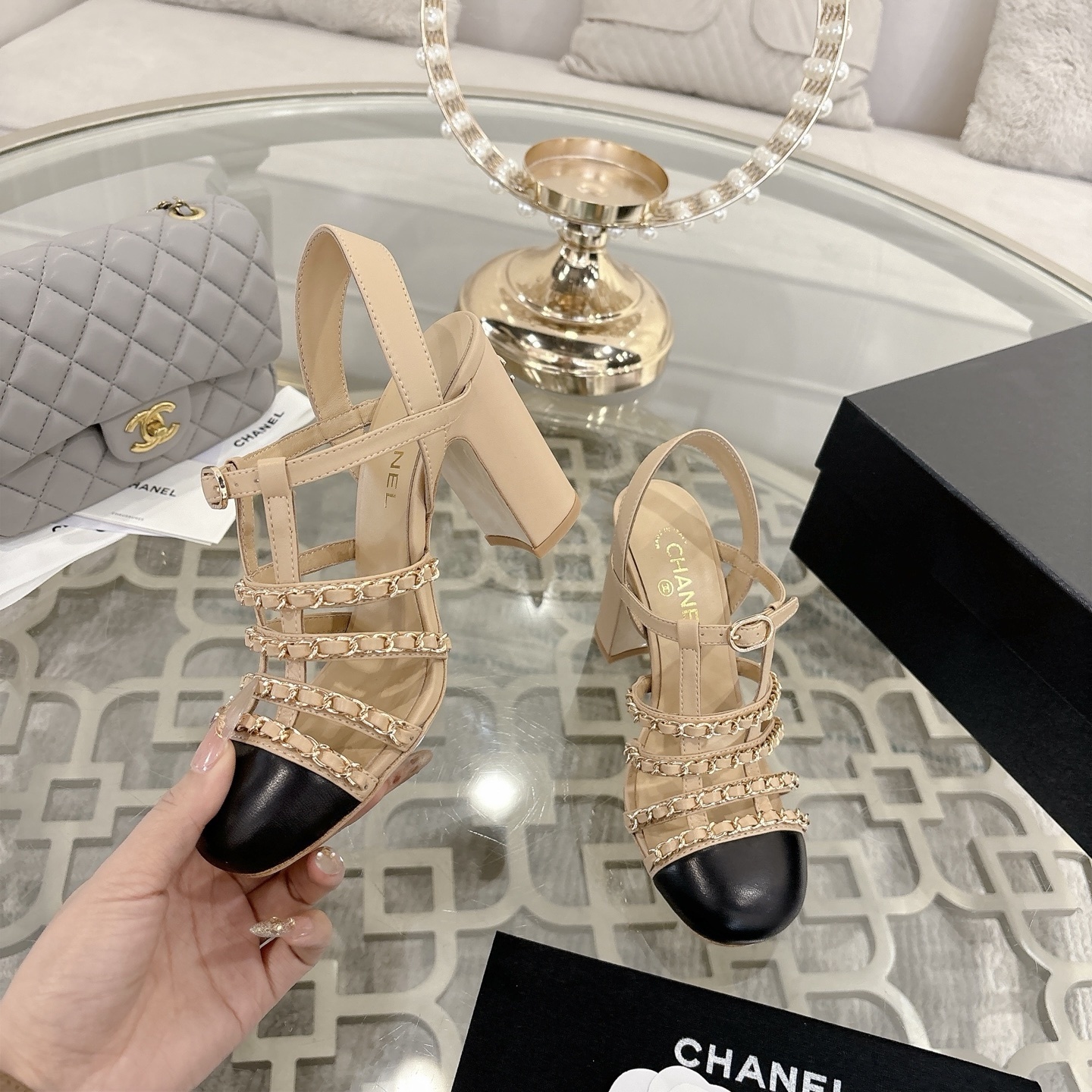 Luxury Chanel beige leather block heel sandals with black cap toes and gold-tone chain straps displayed on a glass table. - Hình 4