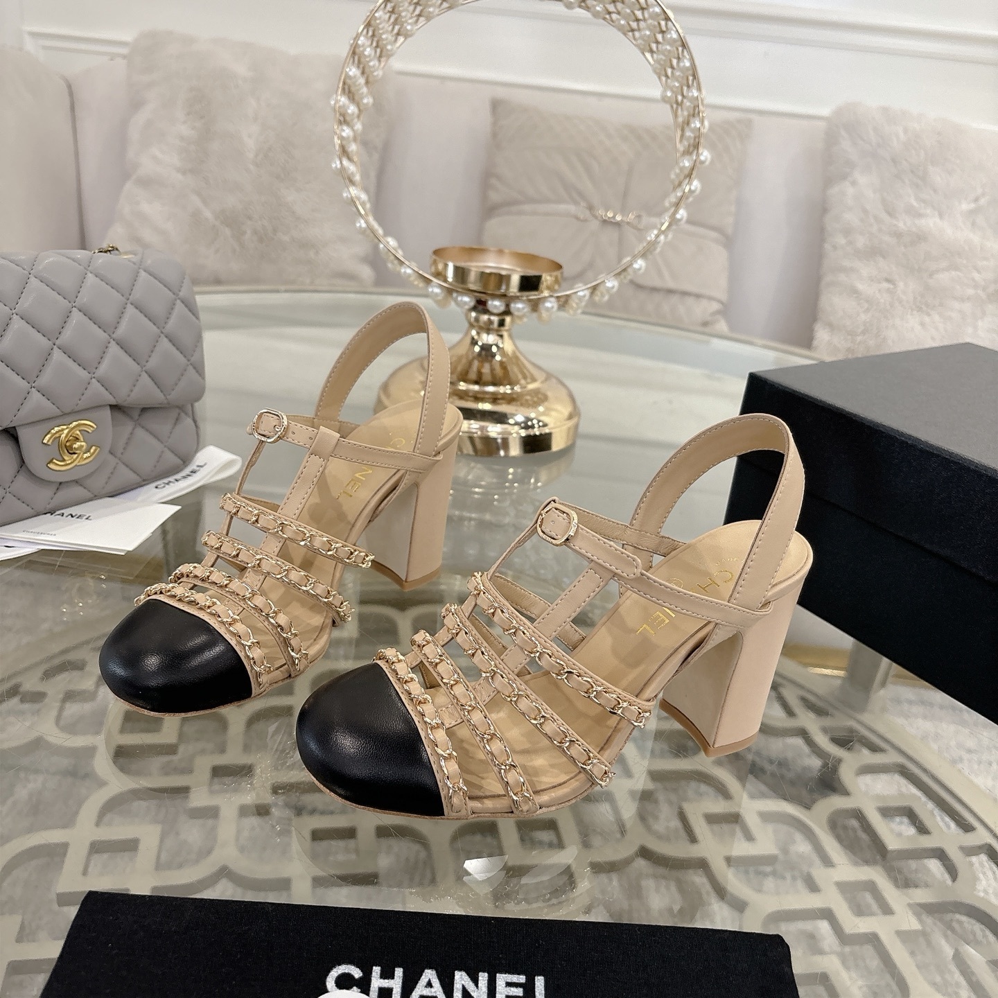 Luxury Chanel beige leather block heel sandals with black cap toes and gold-tone chain straps displayed on a glass table. - Hình 3