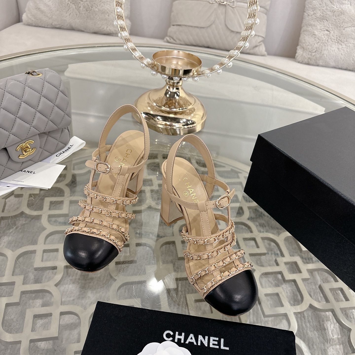 Luxury Chanel beige leather block heel sandals with black cap toes and gold-tone chain straps displayed on a glass table. - Hình 2