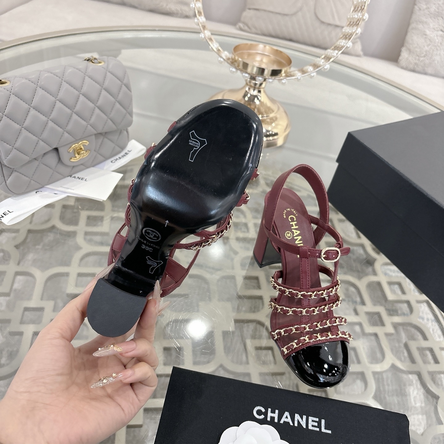 A pair of Chanel burgundy leather block heel sandals with gold chain straps and black patent cap toe displayed on a glass table. - Hình 9