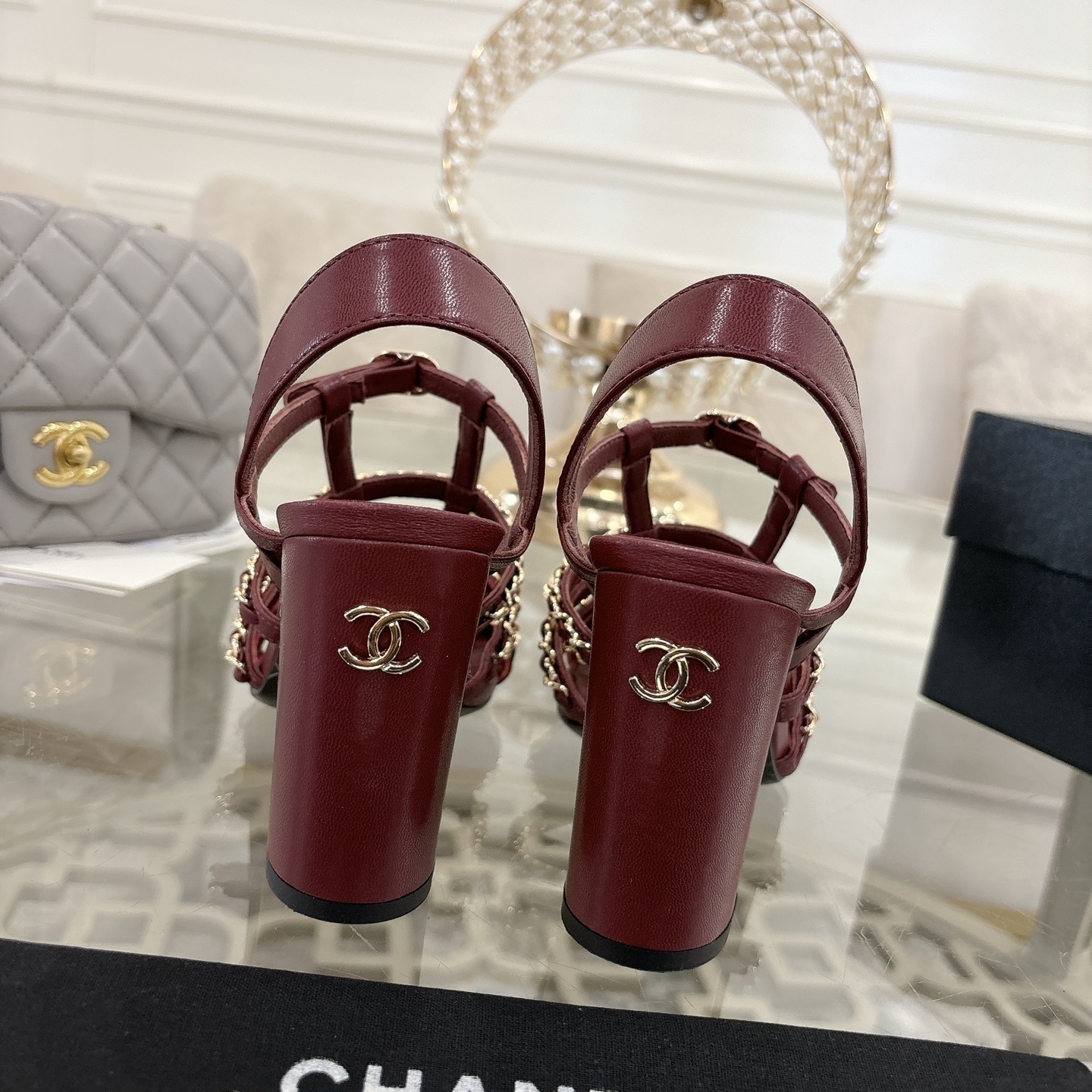 A pair of Chanel burgundy leather block heel sandals with gold chain straps and black patent cap toe displayed on a glass table. - Hình 8