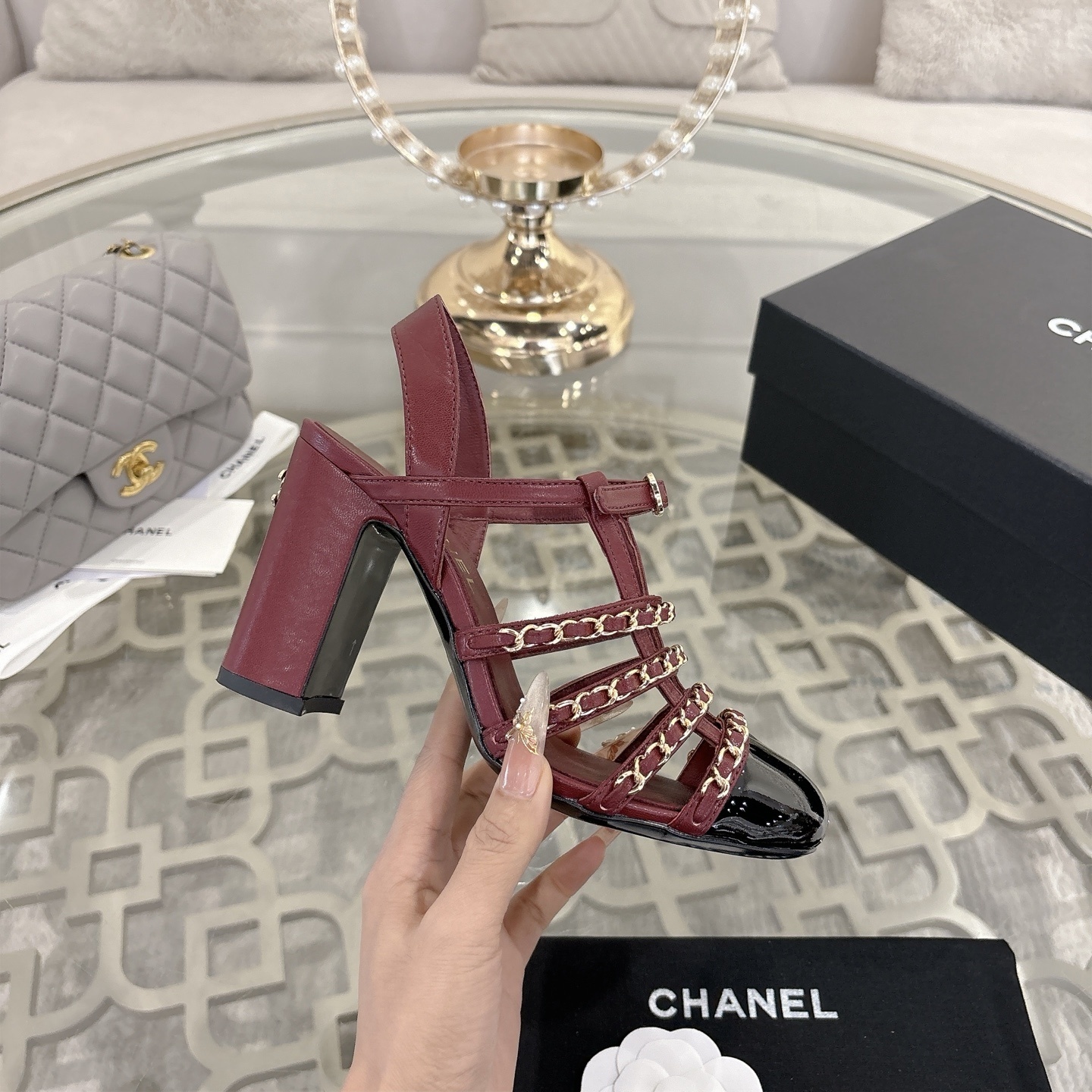 A pair of Chanel burgundy leather block heel sandals with gold chain straps and black patent cap toe displayed on a glass table. - Hình 5