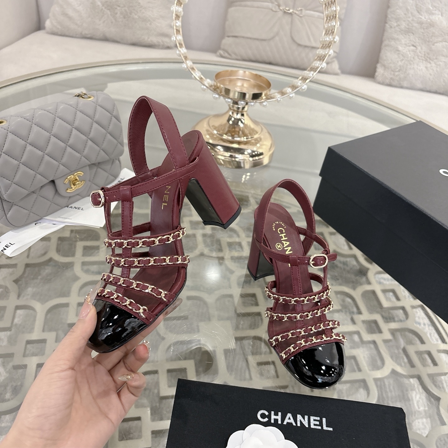 A pair of Chanel burgundy leather block heel sandals with gold chain straps and black patent cap toe displayed on a glass table. - Hình 4